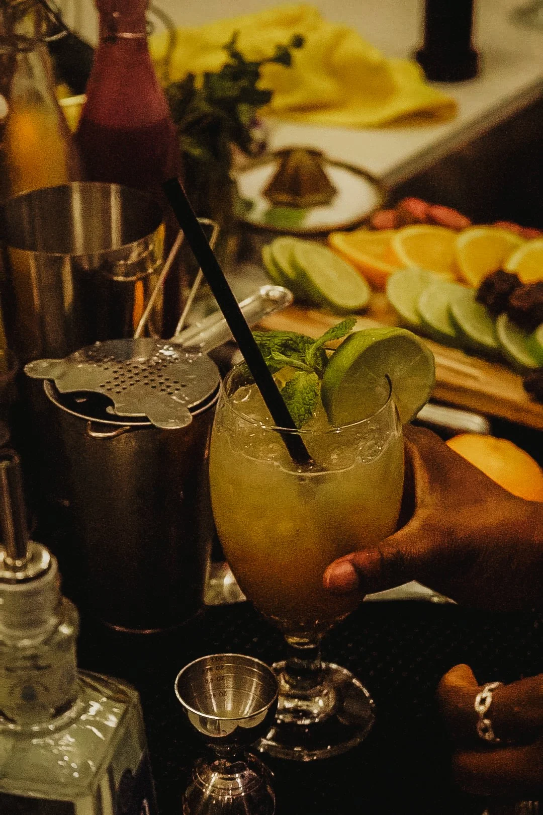 A hand holding a tall glass of cocktail with lime and mint garnish, on a bar counter with various fruits and bar tools in the background.