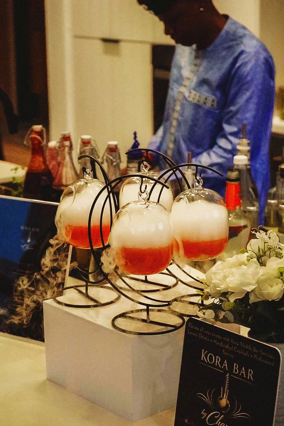Four glasses of Kora Bar signature cocktail with orange hue and foam on top, on a stand, with bottles of various flavors in the background, at a beverage station with flowers and a sign.