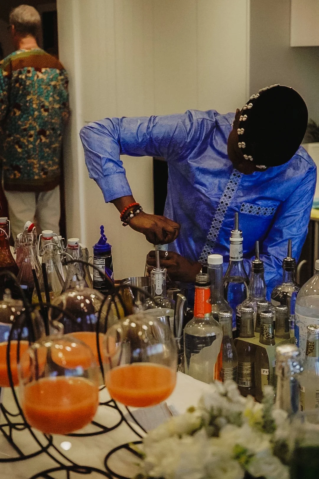 A person in a blue traditional outfit preparing drinks at a bar with various bottles and glasses, and another person in the background.
