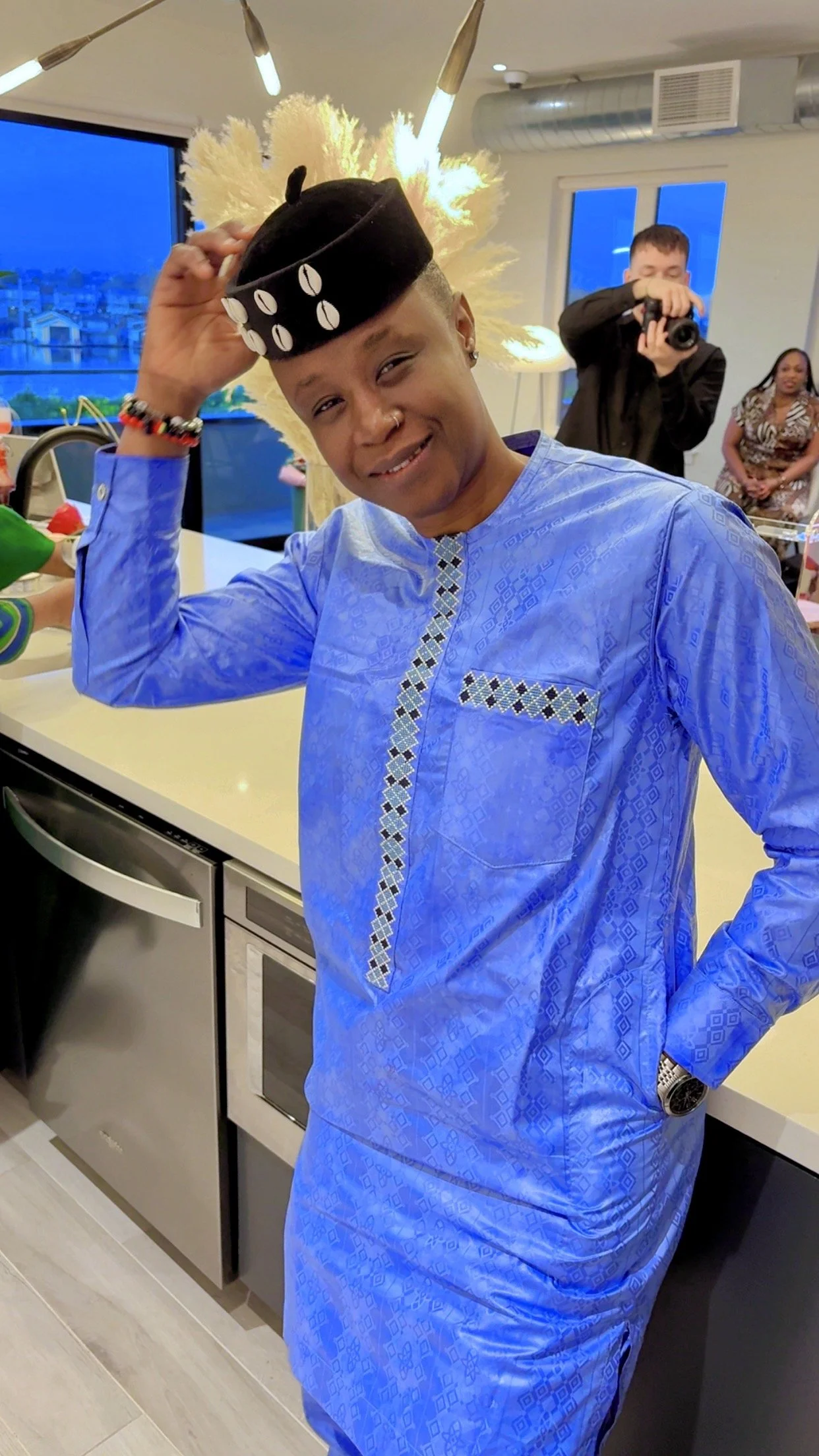 A person wearing a blue traditional African outfit and a black hat with white decorations, standing in a modern kitchen with a camera person and other people in the background.