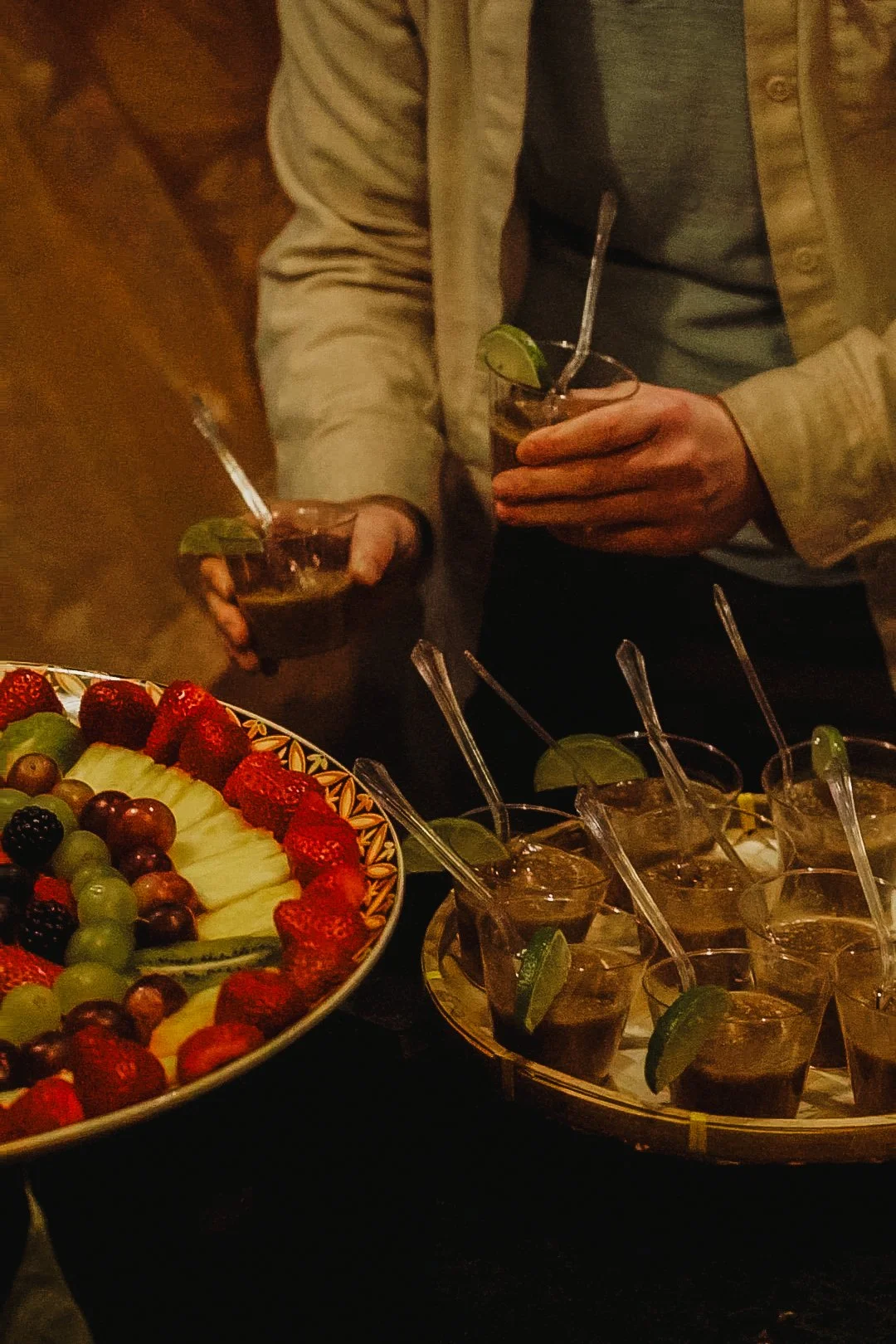 People holding drinks with lime wedges near a fruit platter with strawberries, grapes, and melon slices.