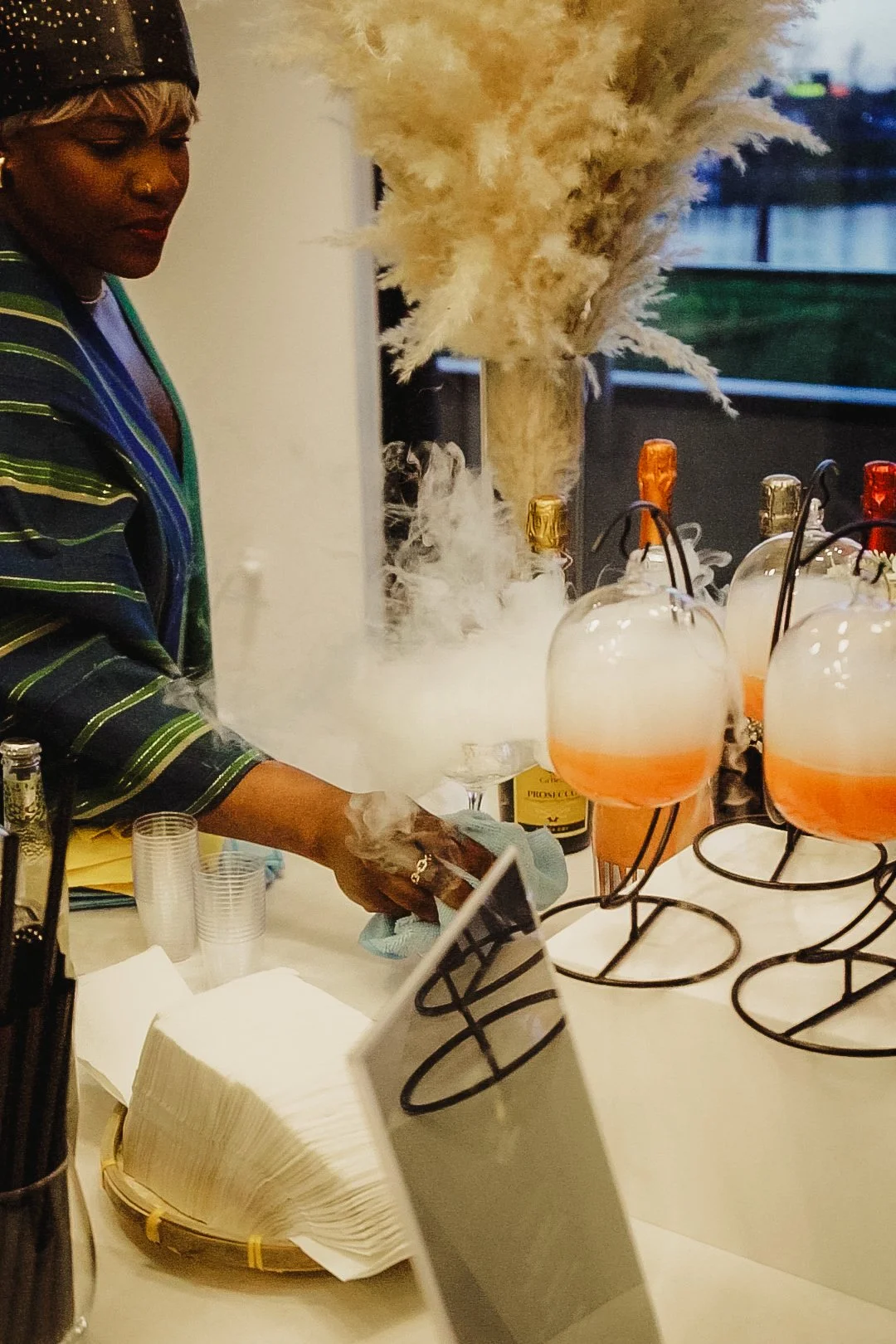 A bartender pouring liquid into glasses with dry ice fog and large decorative pampas grass in the background.