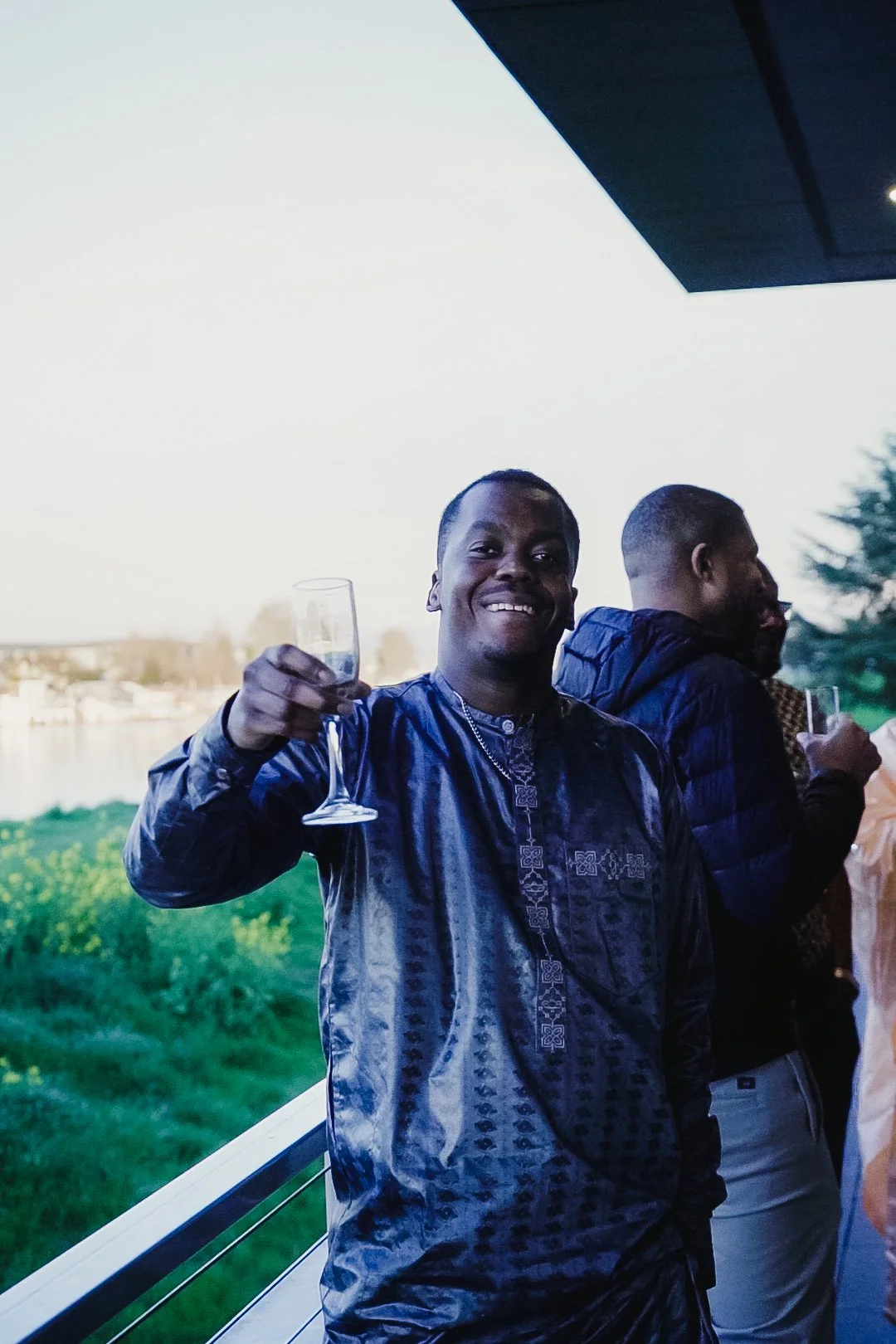 A man smiling and holding a glass of champagne at an outdoor event, with two other men in the background, near a body of water and greenery.