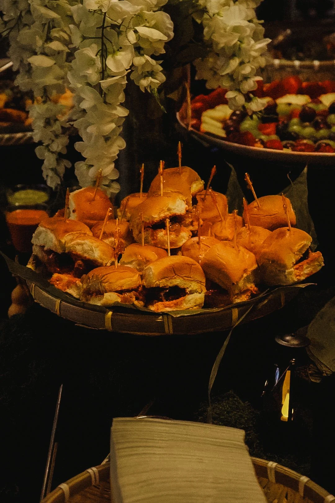 Tray of small sandwiches or sliders with toothpicks on a dark serving tray, with a backdrop of white floral decorations and a bowl of vegetable or fruit salad.