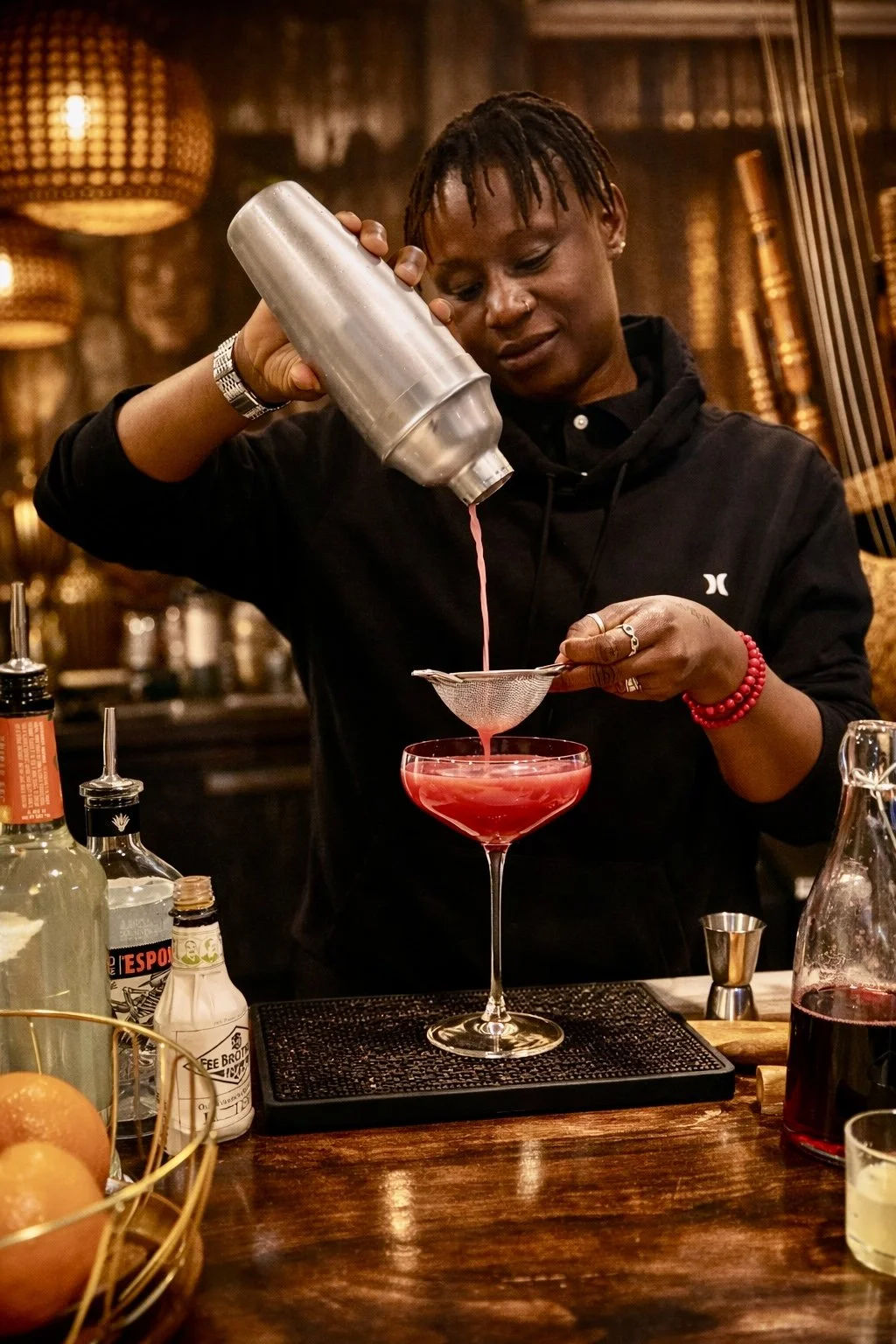 A bartender pours a pink cocktail through a strainer into a glass at a bar with warm lighting and bottles in the background.