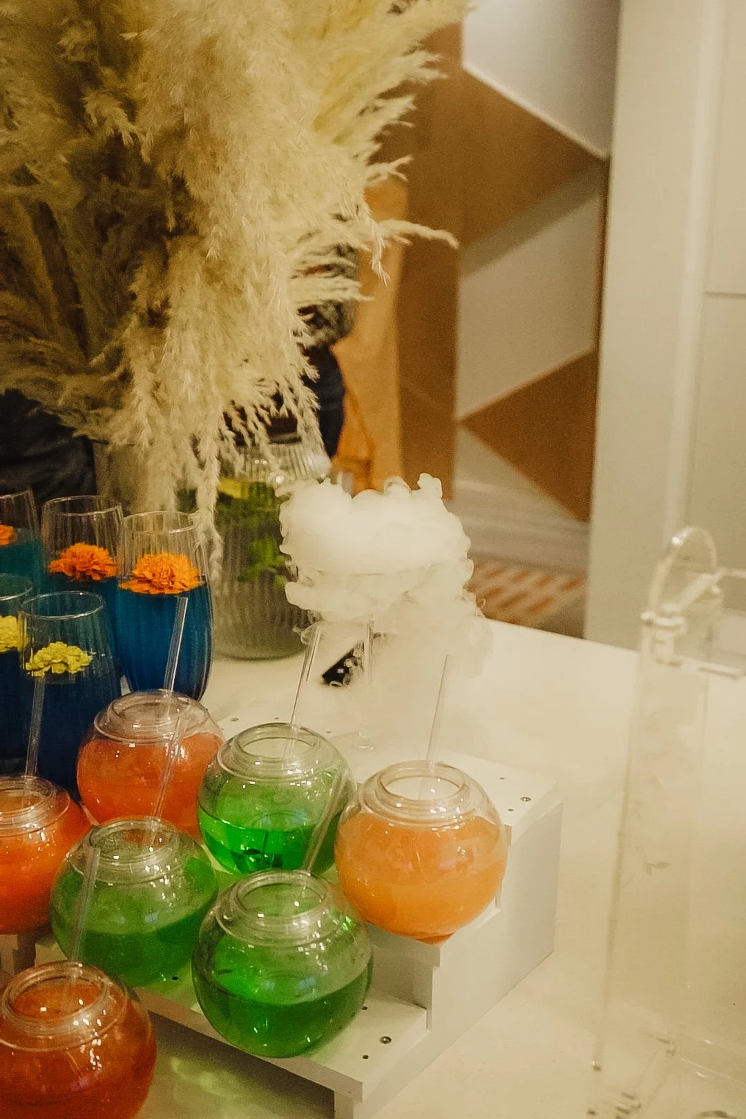 Colorful drinks in glasses, some with straws, arranged on a white table, with dry ice creating fog in the background. Tall glasses with orange and blue liquids, smaller glasses with green and orange liquids.