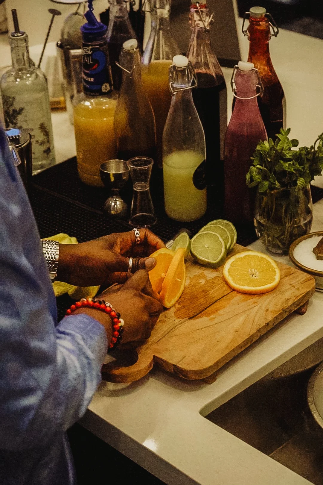 A person slicing lemons, limes, and oranges on a wooden cutting board surrounded by bottles of fruit juices and a potted plant on a kitchen counter.
