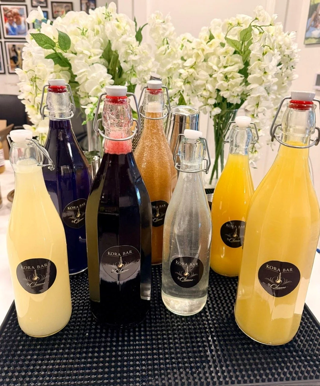 Seven glass bottles with labels reading 'Kora Bar,' filled with various colored liquids, on a textured black surface, with a background of white flowers and framed pictures.