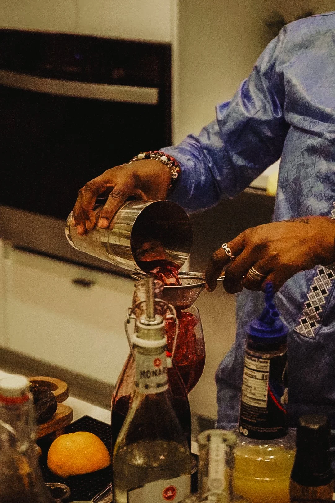 Person pouring red liquid from a shaker into a glass with various bottles, a lemon half, and bar tools on the counter.