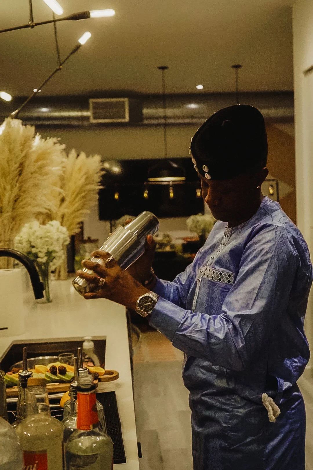A person dressed in a traditional blue African attire, wearing jewelry and a watch, is holding a silver container in a modern kitchen with bottles, fruits, and flowers in the background.