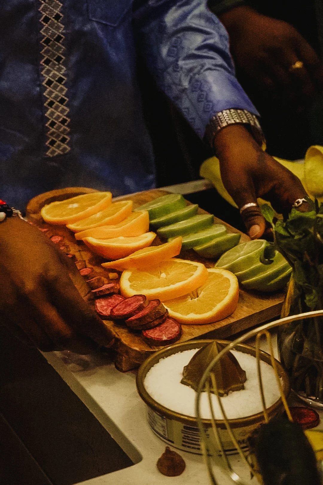 Person slicing oranges, cucumbers, and salami on a wooden cutting board, with a bowl of salt and a knife nearby.