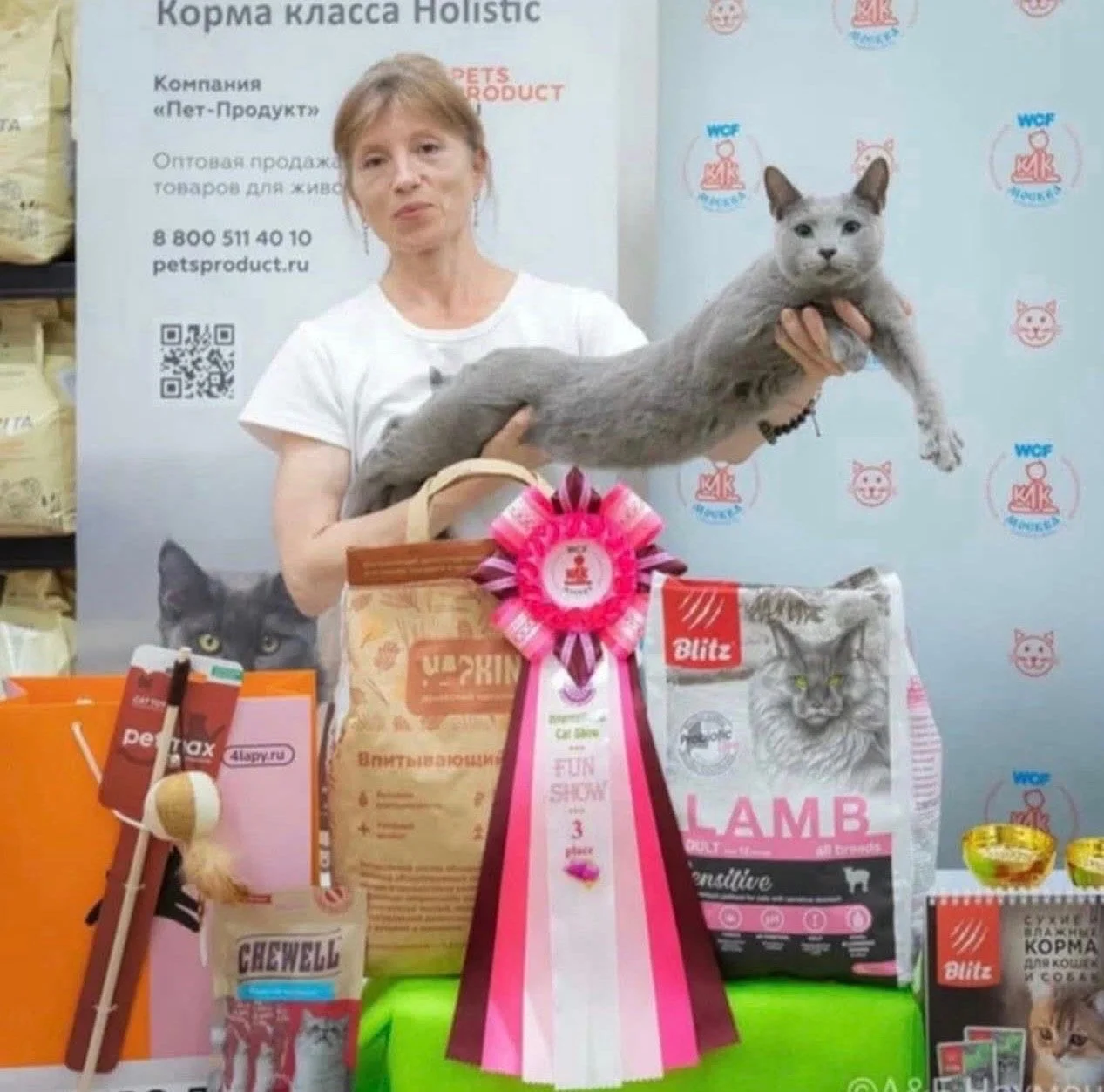 Woman holding a large gray cat at a pet products event with pet food and supplies displayed on the table.