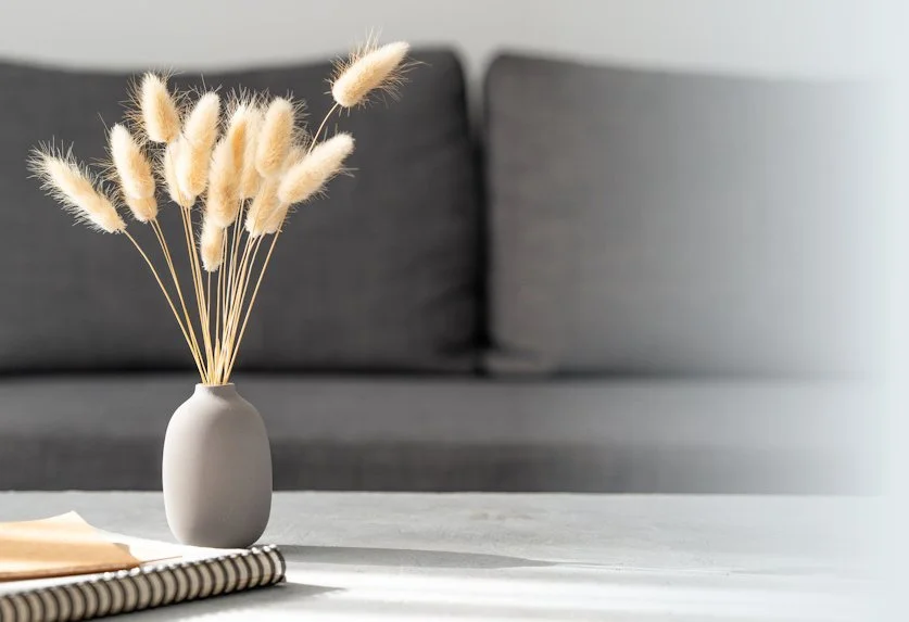 A white vase with dried wheat stalks placed on a table in front of a gray sofa.