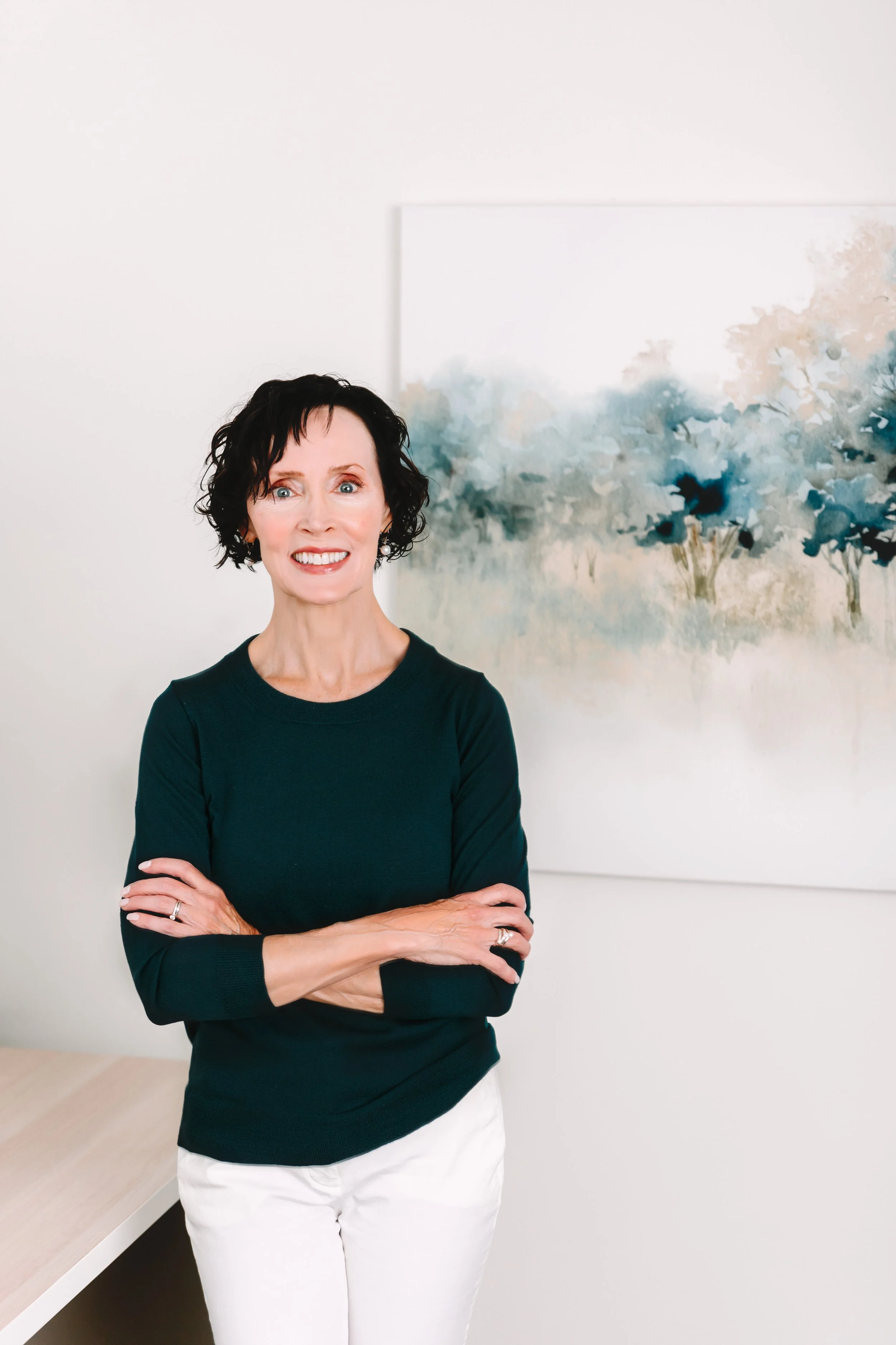 A woman with short dark hair, wearing a dark teal long-sleeve top and white pants, standing with her arms crossed in front of a white wall with a framed abstract painting of blue and beige trees.