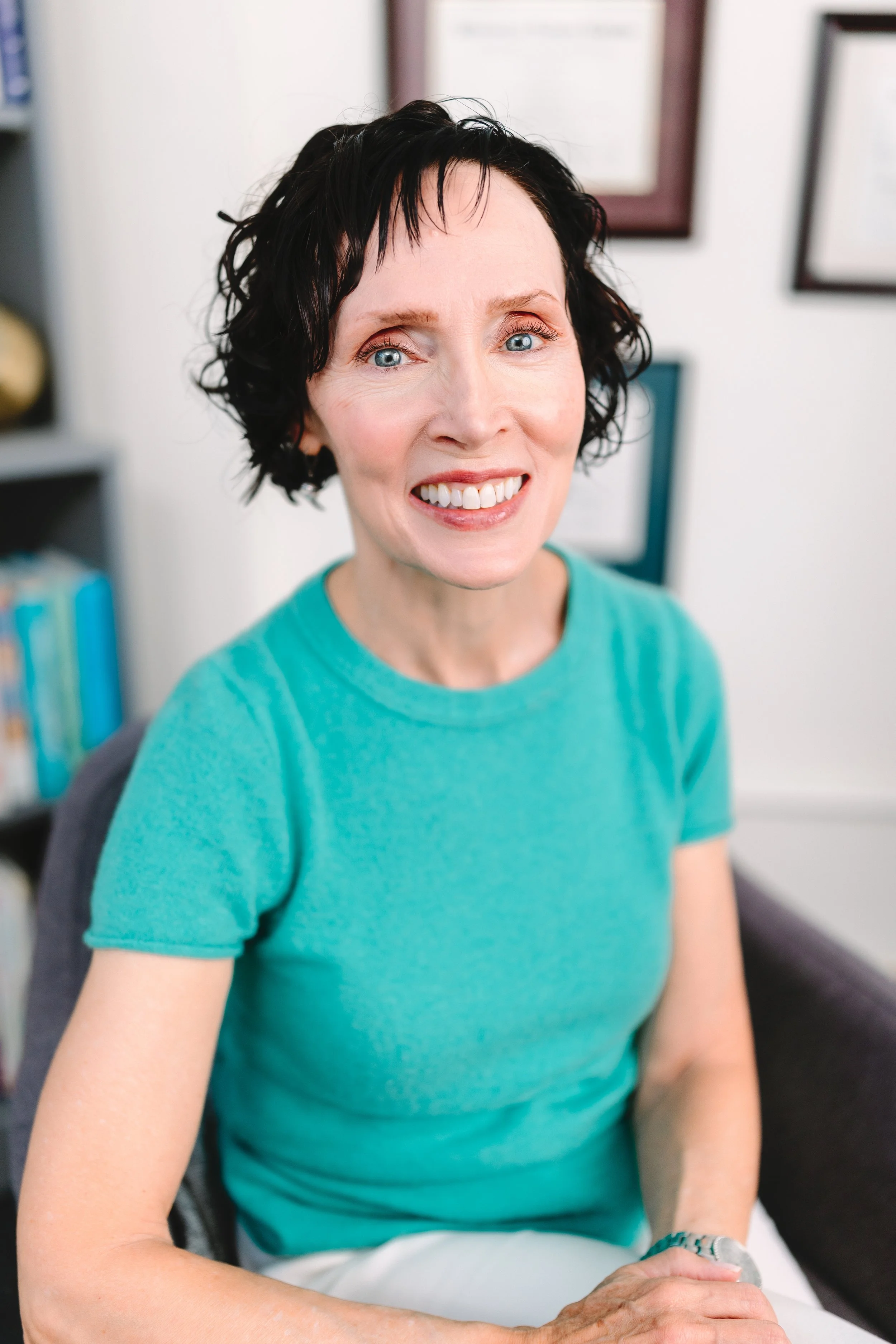 A woman with short black hair, wearing a turquoise shirt, smiling and sitting in an indoor setting with framed pictures on the wall behind her.