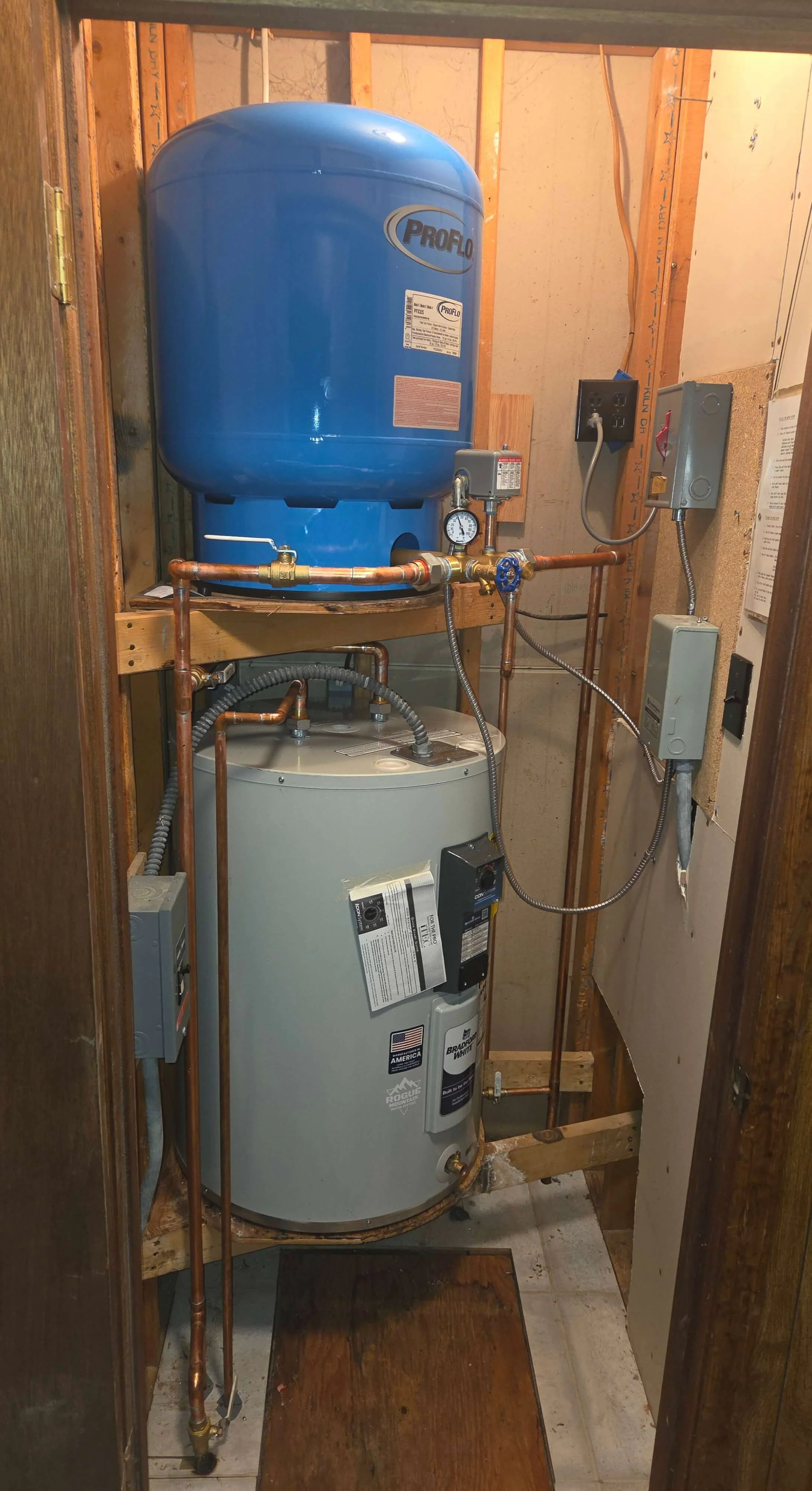 A water heater and expansion tank installed in a small utility closet with exposed wooden framing, electrical wiring, and copper plumbing pipes.
