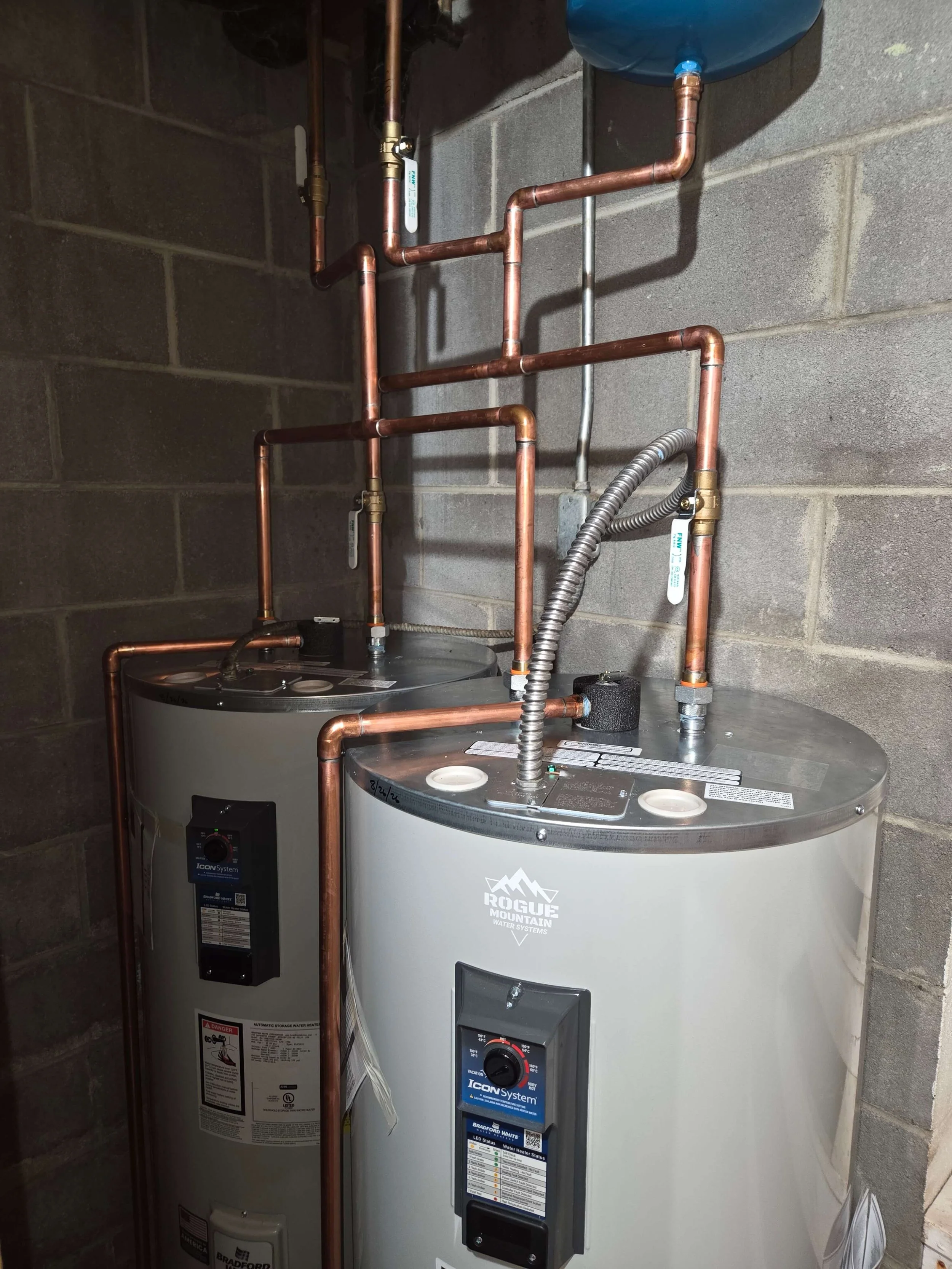 Two water heaters with copper pipes and gauges in a basement against a concrete block wall.