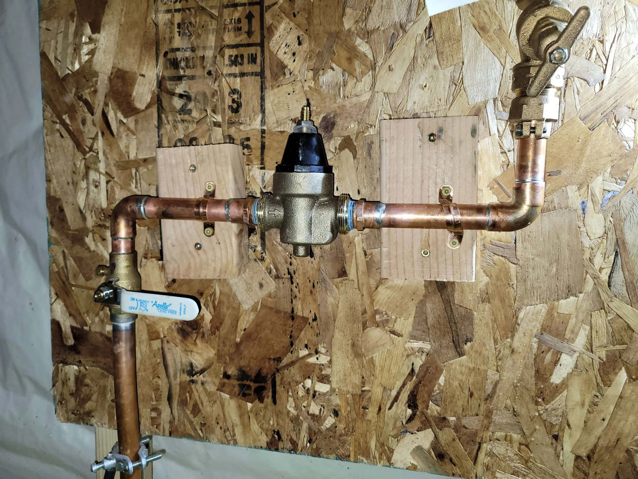 Copper piping system with a pressure valve and shut-off valve on a plywood wall.