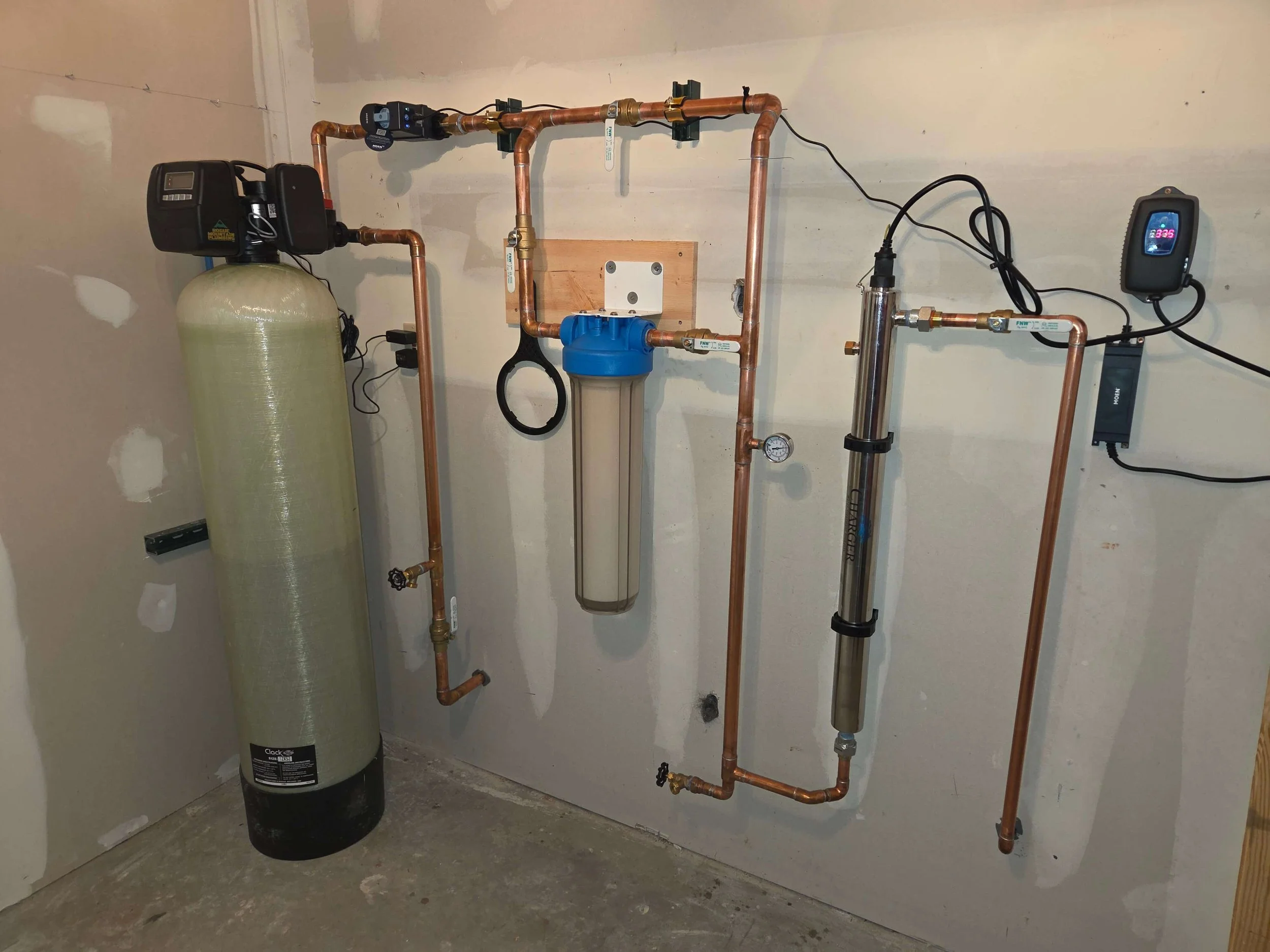 A water treatment system with copper pipes, a water filter, and electronic control devices installed on a drywall wall in a basement or utility room.