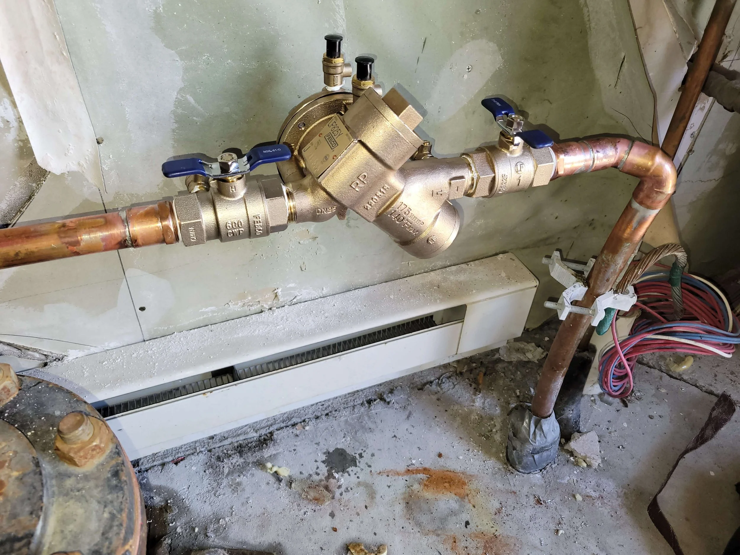 Plumbing pipes and valves in a utility room, featuring copper pipes connected to brass valves, with blue handles for water flow control. Wall and floor show signs of construction or maintenance work.