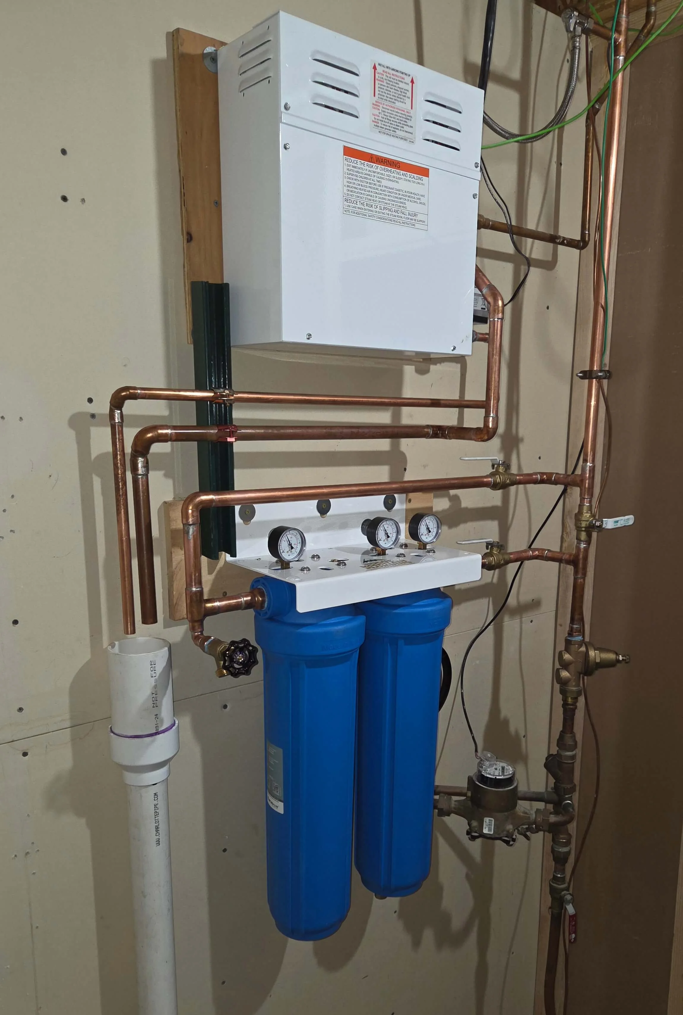 Whole house water filtration system with copper pipes, gauges, filters, water meter, and electrical box mounted on a wall in a utility room.