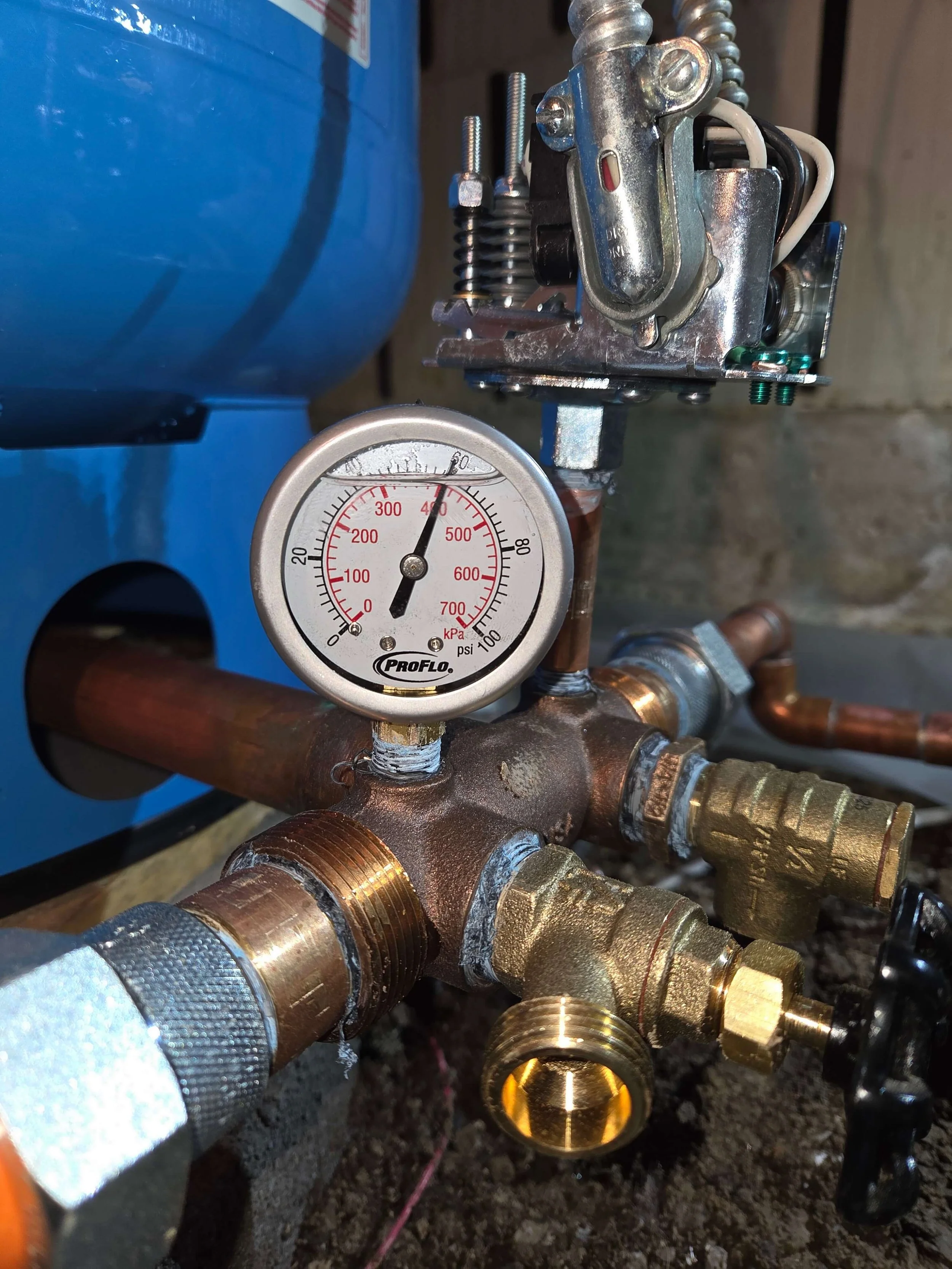 Close-up of plumbing system with brass fittings, copper pipes, and a pressure gauge reading approximately 60 PSI, mounted on a copper pipe with a water tank in the background.