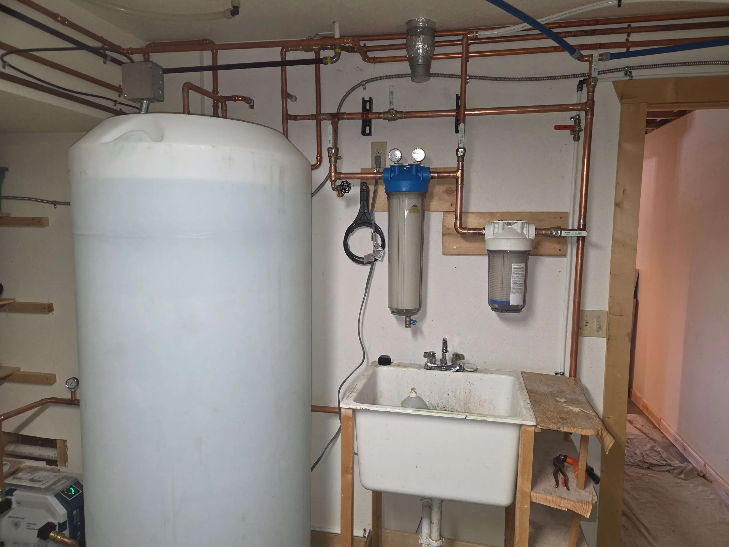 A utility room with a water filtration system, a large water storage tank, and a utility sink with a wooden countertop. Copper pipes and water filters are installed along the wall.