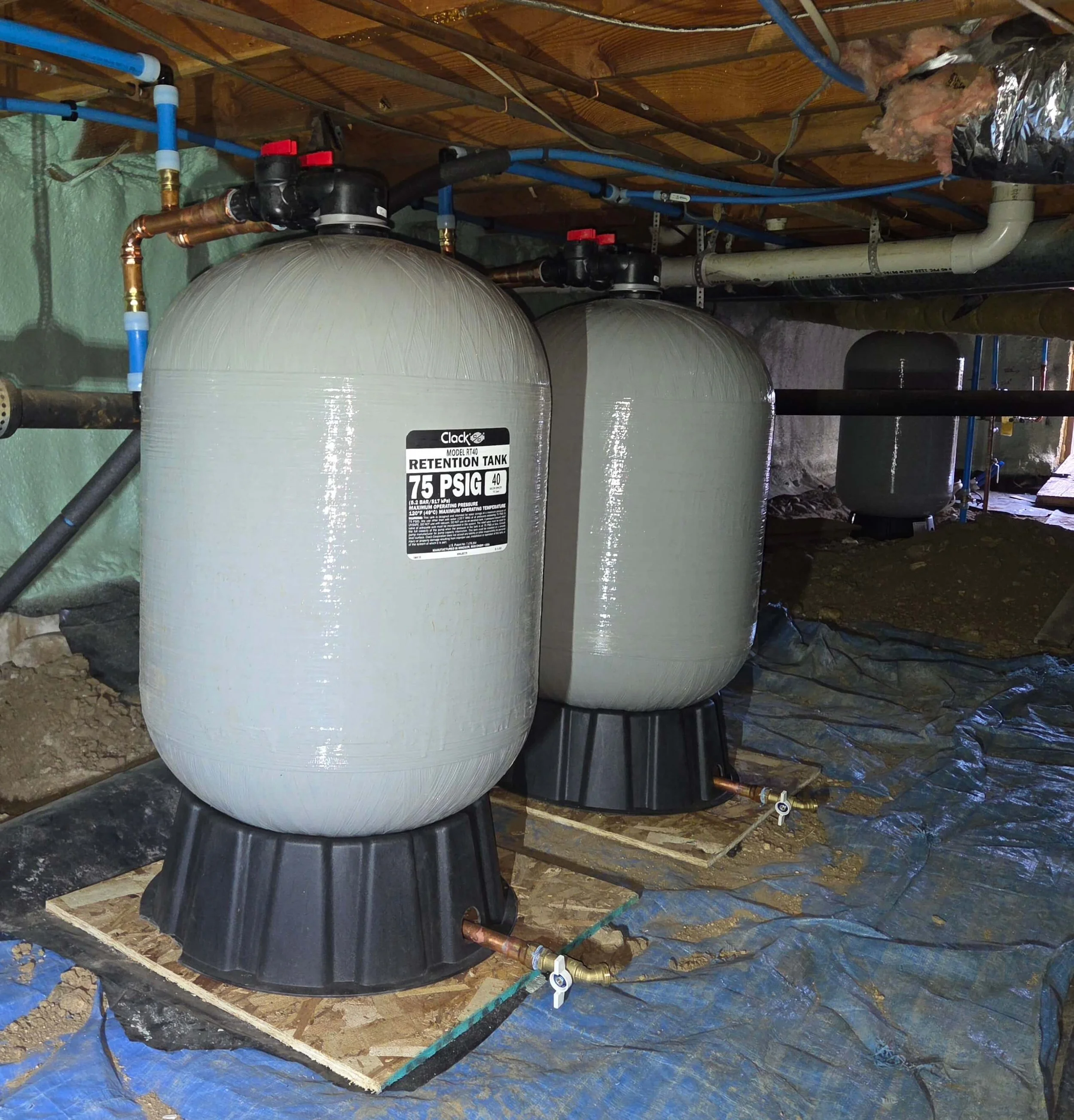 Two water tanks installed in a basement with plumbing connections and a sump pump on a wooden platform, covered with a tarp on the floor.