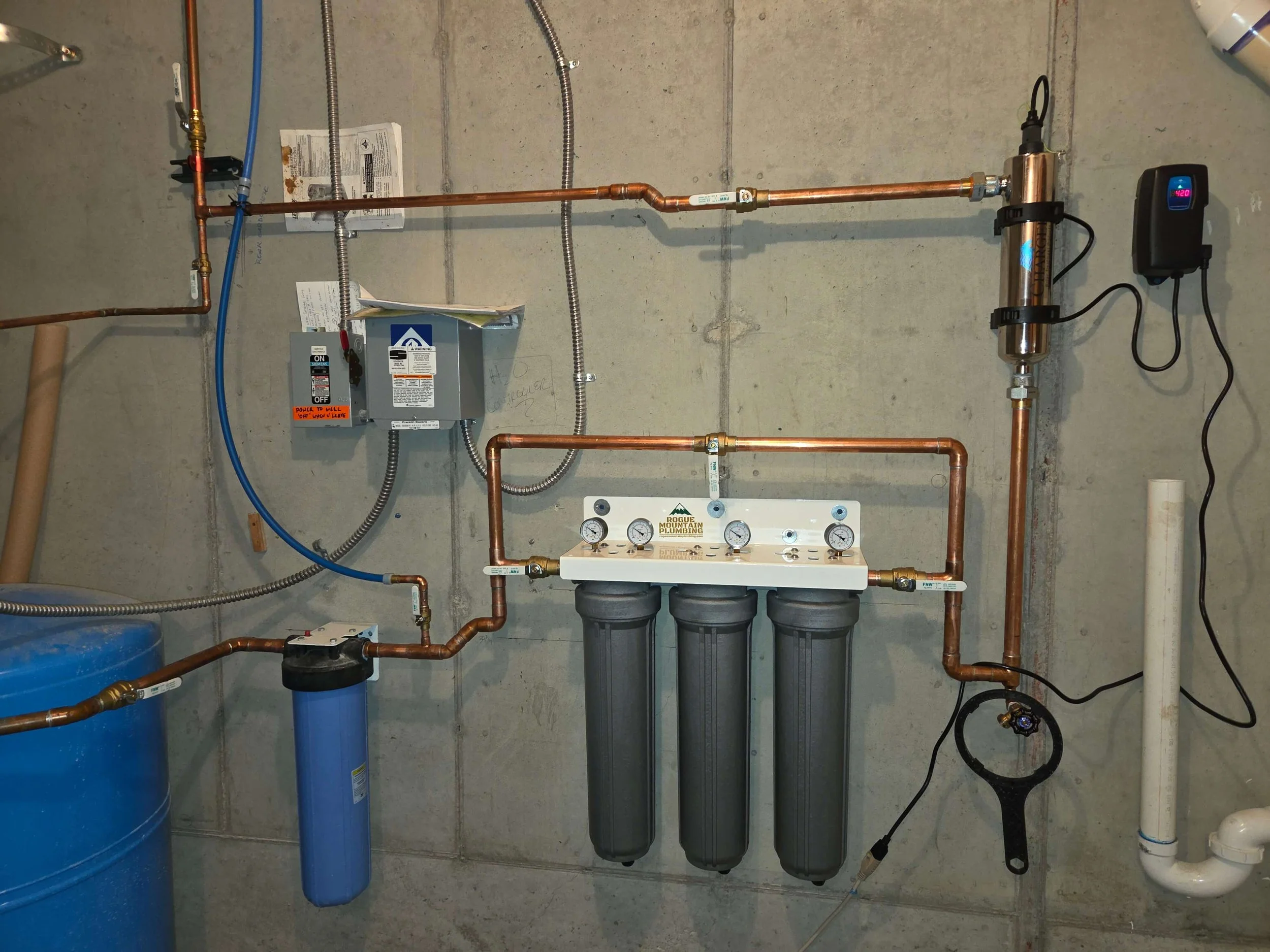 A water filtration system with copper pipes, filters, gauges, and a control box mounted on a concrete wall.