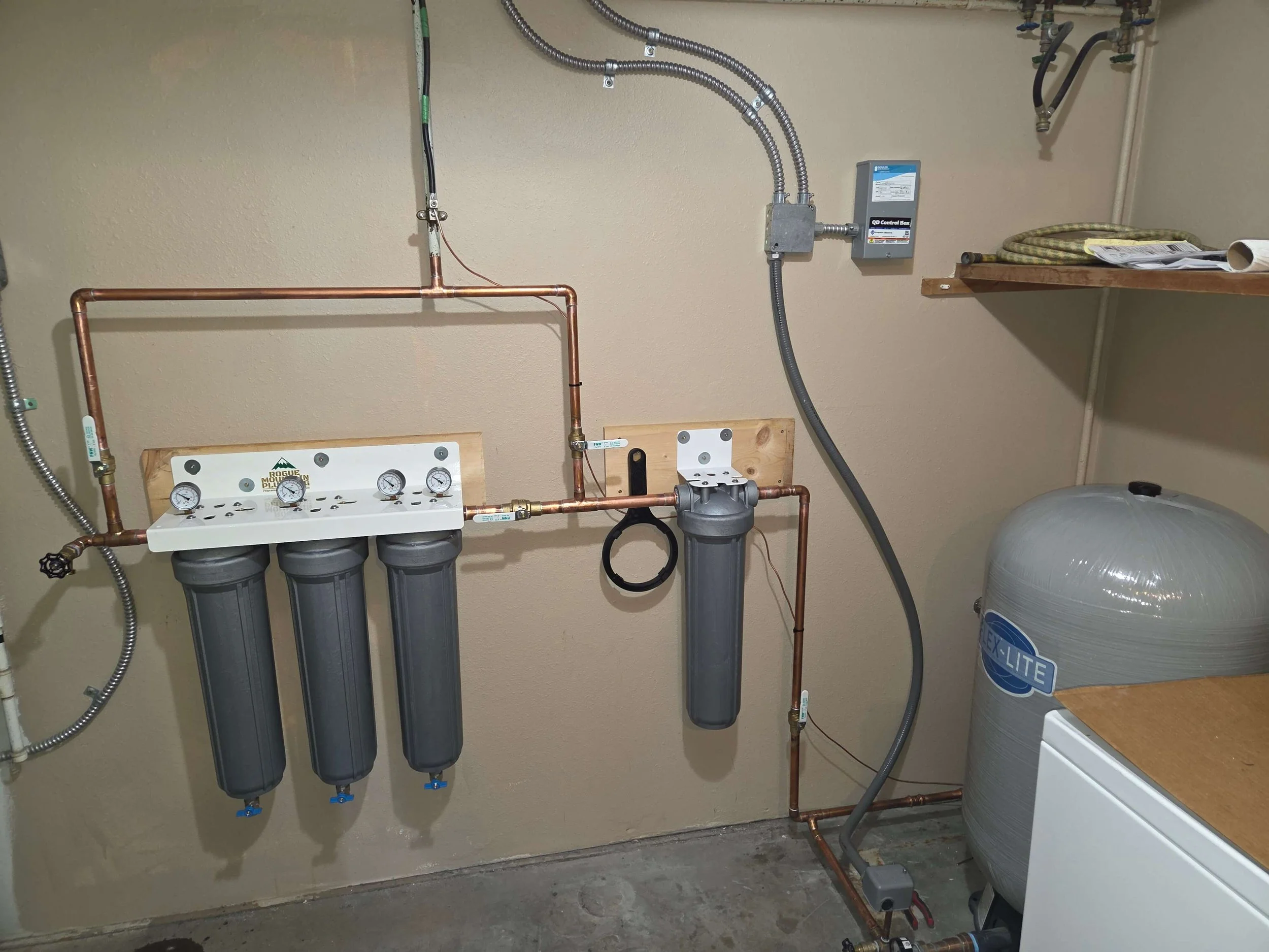 Water filtration system with three black filter canisters, pressure gauges, copper piping, and an additional filter mounted on a beige wall in a utility room.