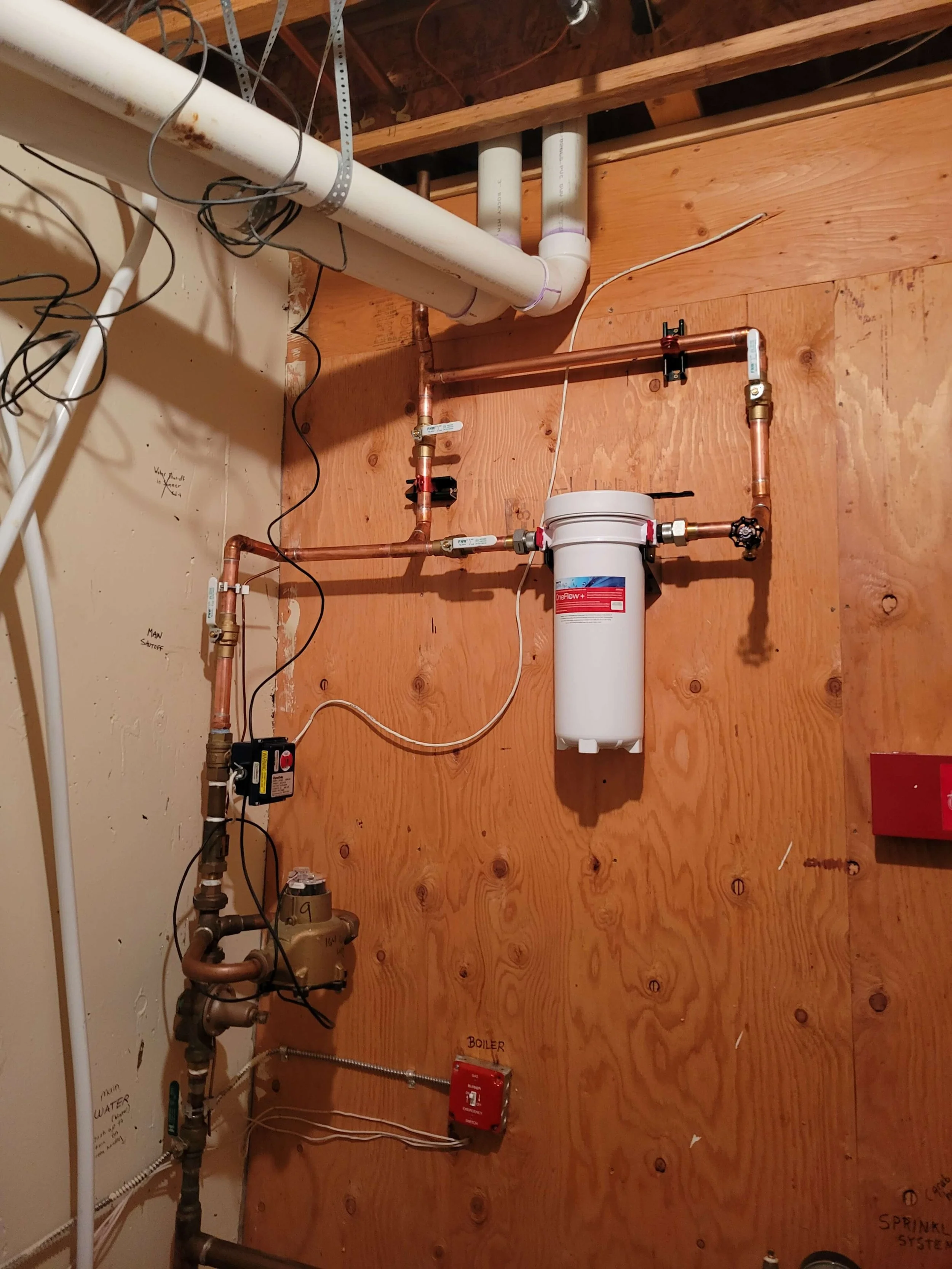 Basement utility room with plumbing pipes, pressure gauge, valves, and a water filtration system mounted on a plywood wall.