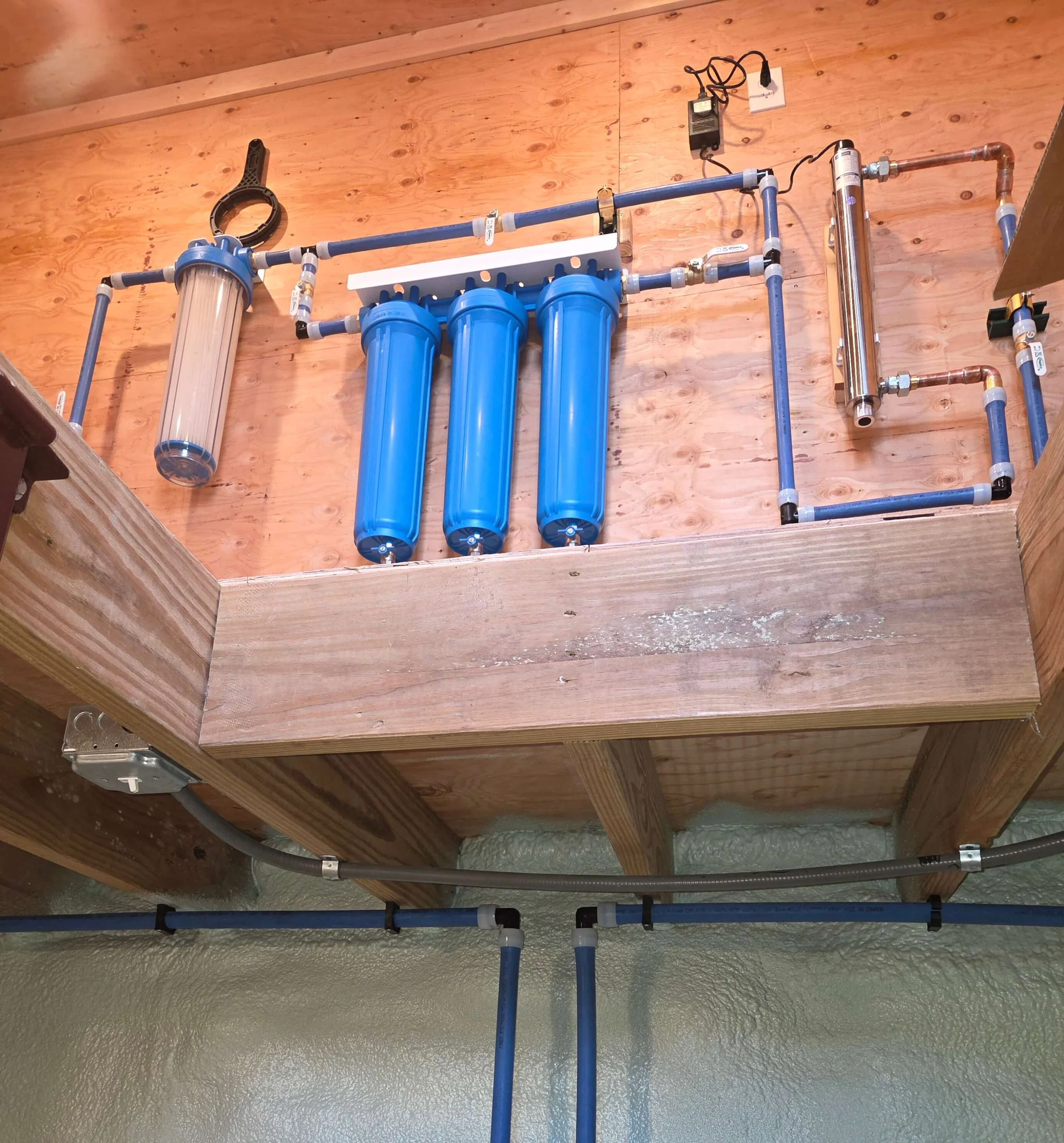 Water filtration system with three large blue filter housings, one clear housing, and connected blue and copper pipes installed on a wooden wall in a construction space.