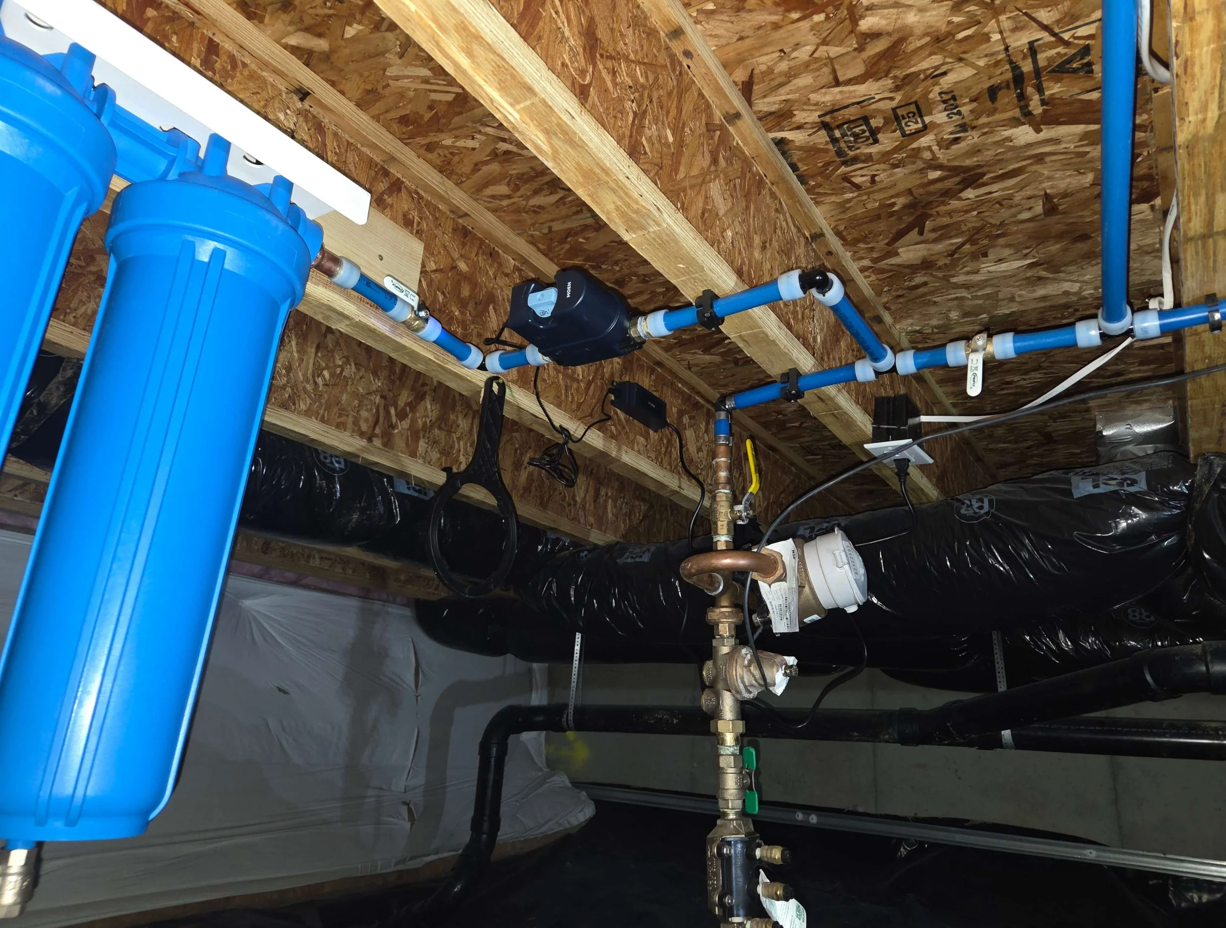 Plumbing system with blue water filter housing, pipes, valves, and black insulation in a wood-framed space.