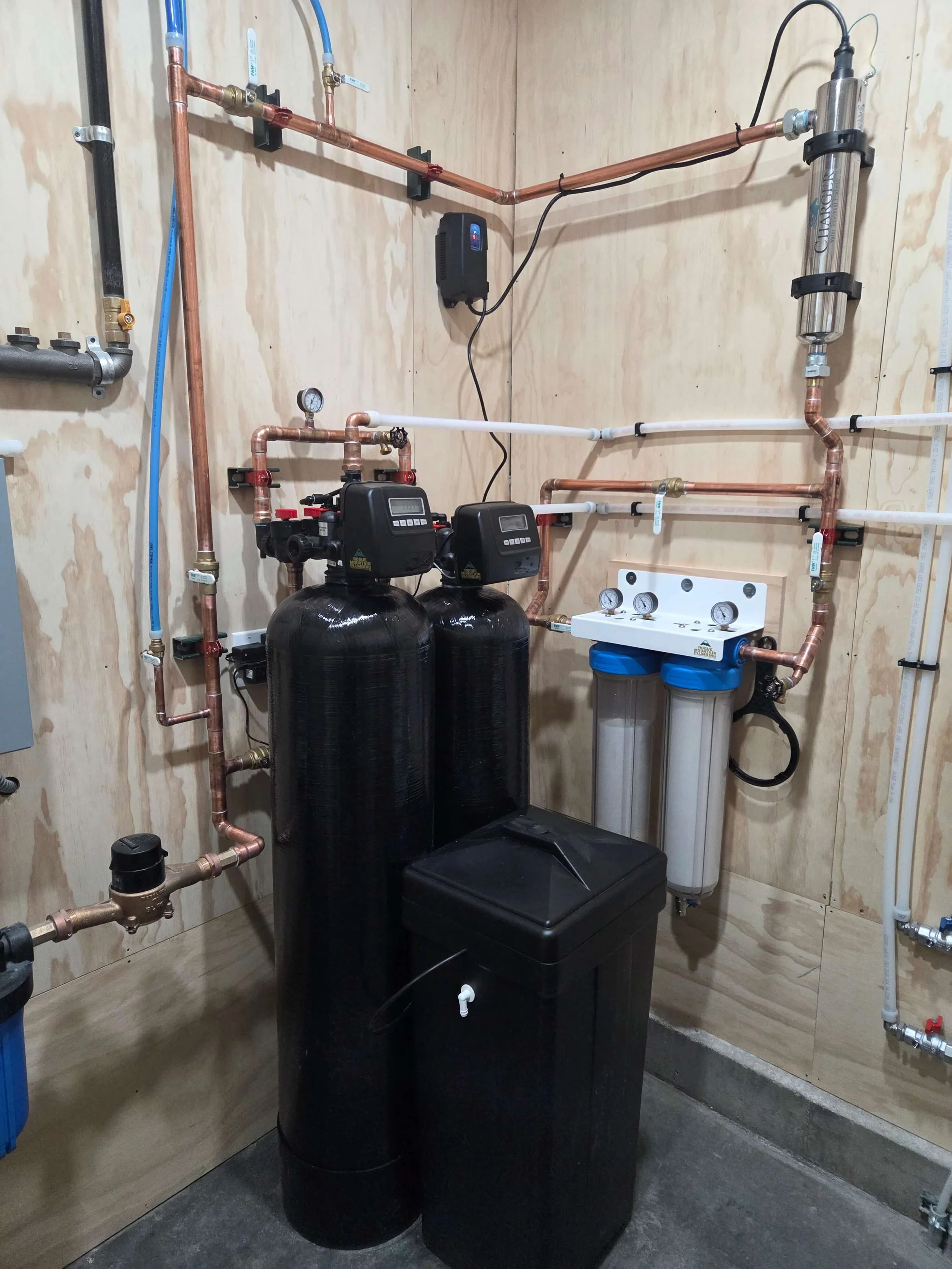 Water filtration system with black tank, pipes, gauges, and filters mounted on wood-paneled wall.