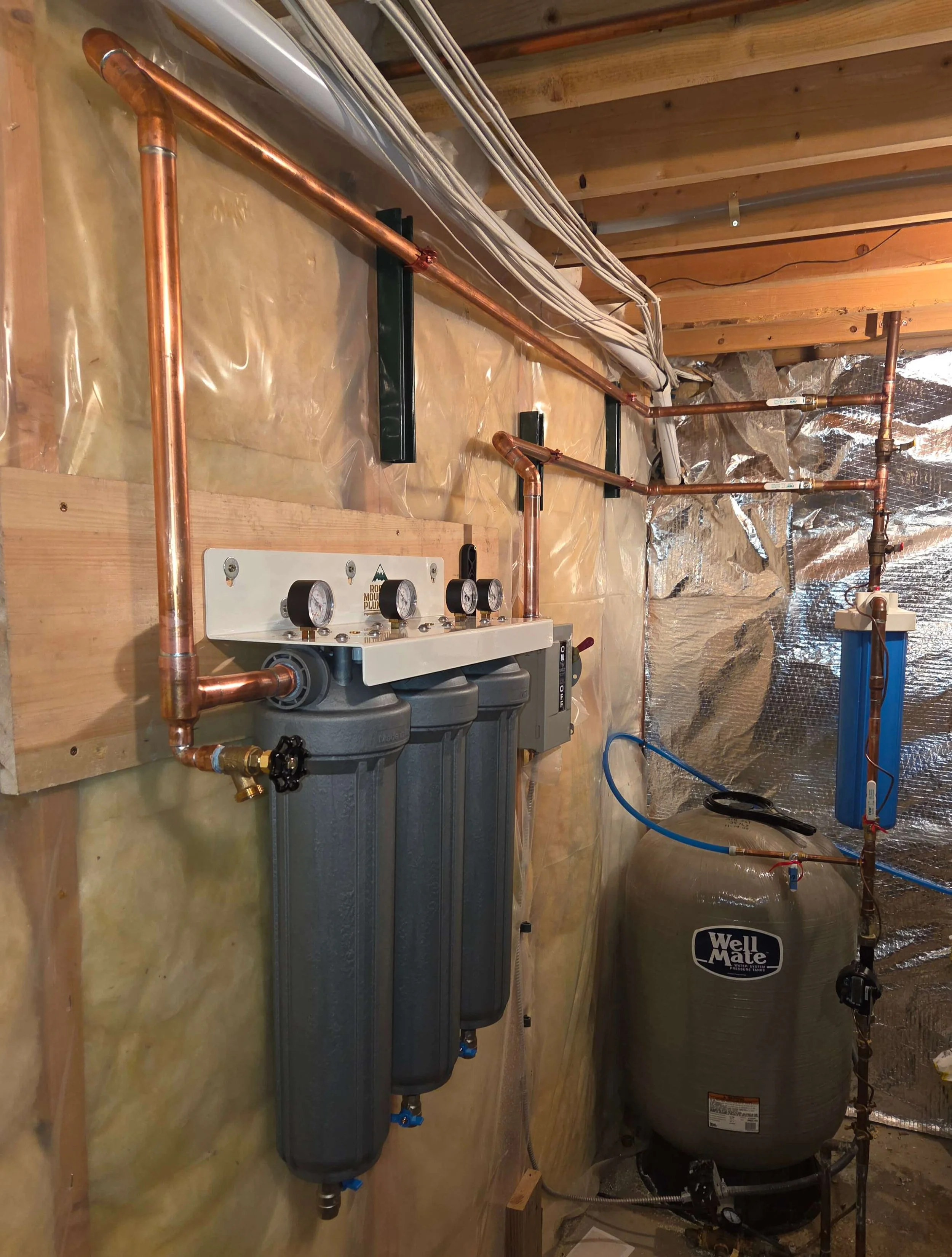 A water filtration system installed on a wall inside a building with copper pipes, gauges, and a large Well Mate pressure tank.