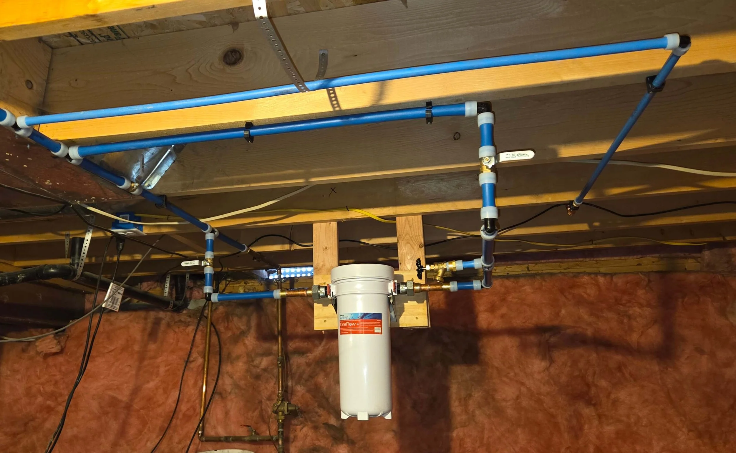 Basement ceiling with blue PEX plumbing pipes and a water filter system, surrounded by insulation and electrical wires.