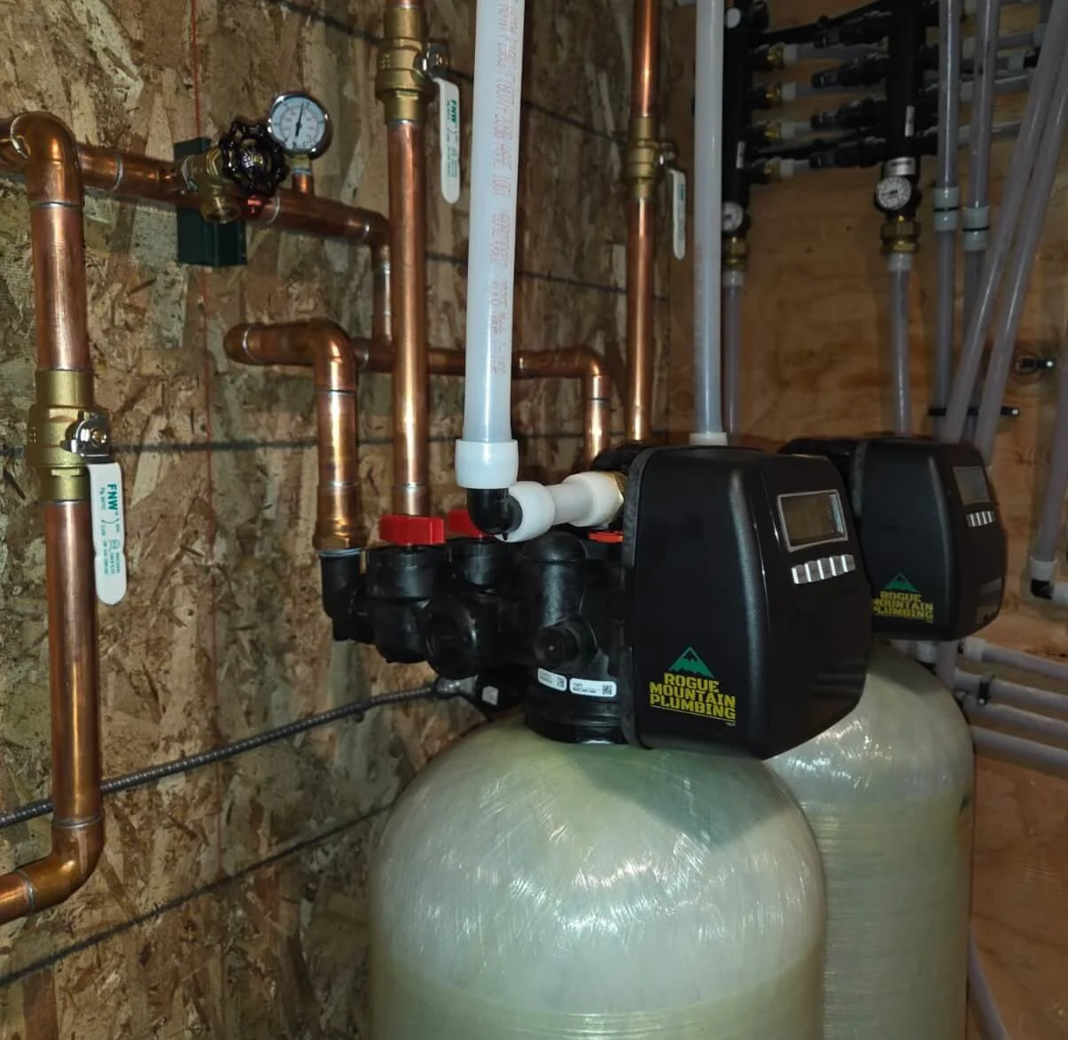 Water filtration system with two large beige tanks, black control valves labeled "Rogue Mountain Plumbing," copper pipes, and white PVC pipes in a wooden utility room.
