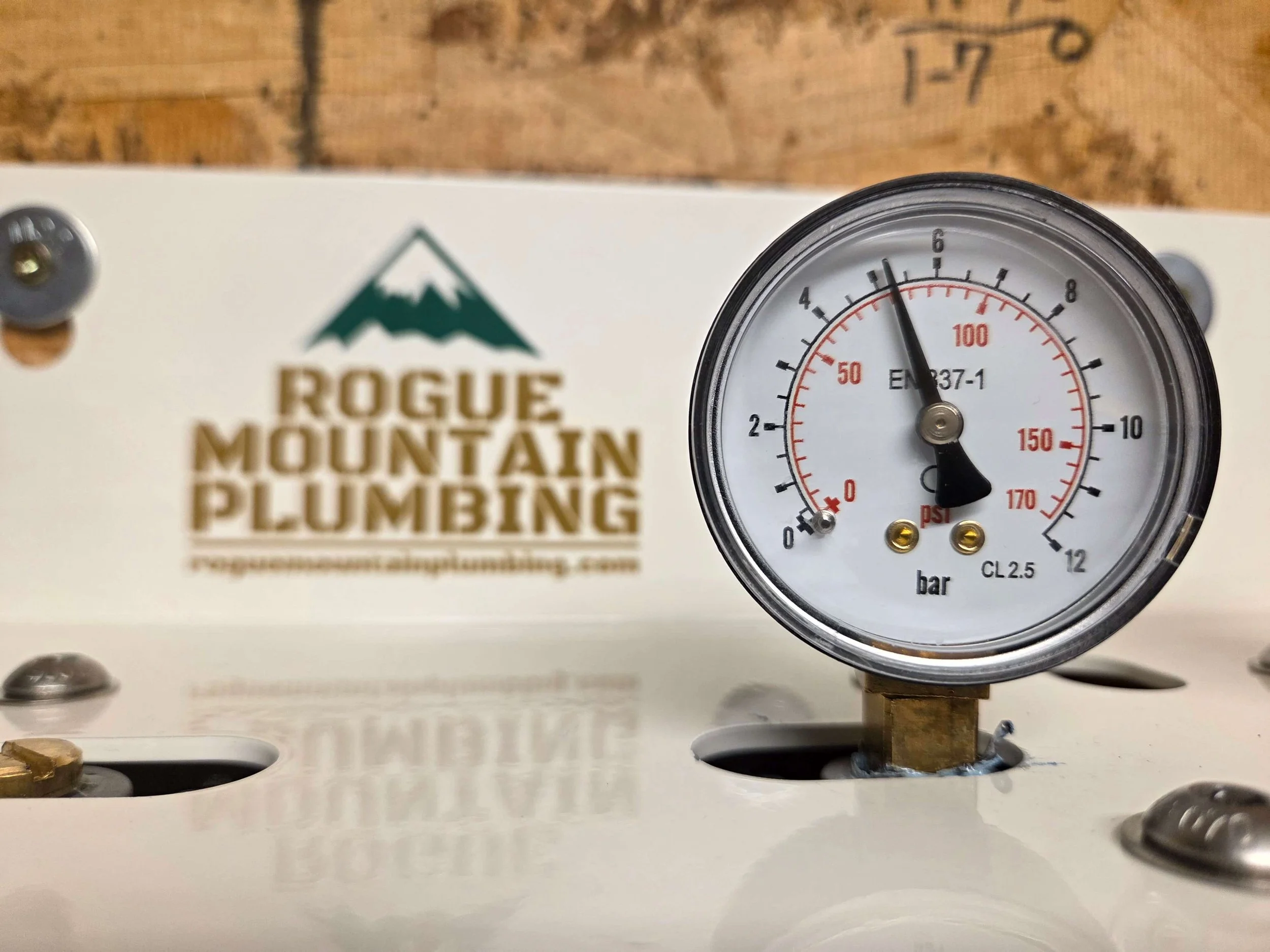 Pressure gauge showing approximately 6 psi pressure, mounted on a piece of plumbing equipment with "Rogue Mountain Plumbing" logo in the background.