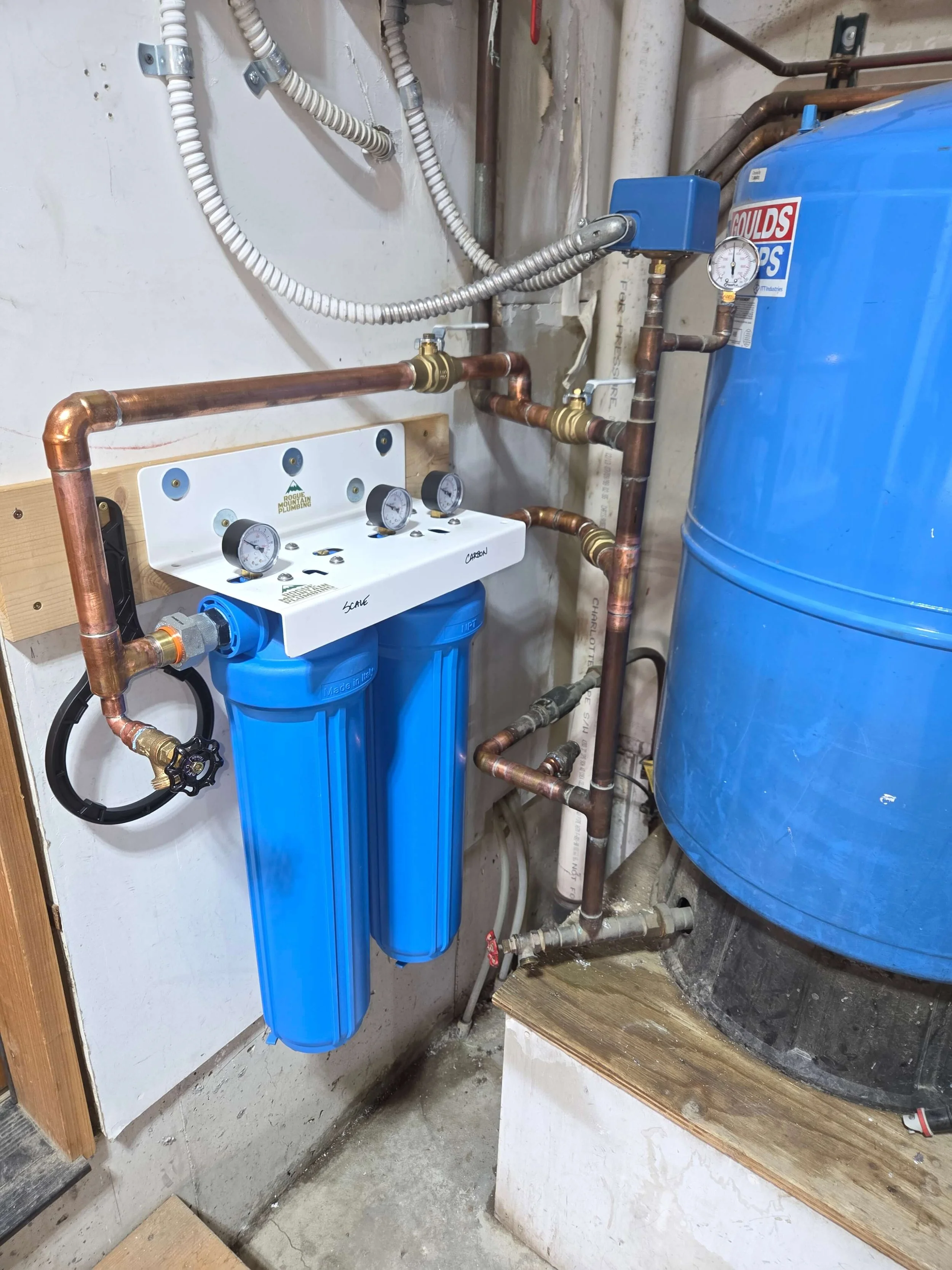 A water filtration system installed next to a large blue tank, with copper pipes, gauges, and filters.