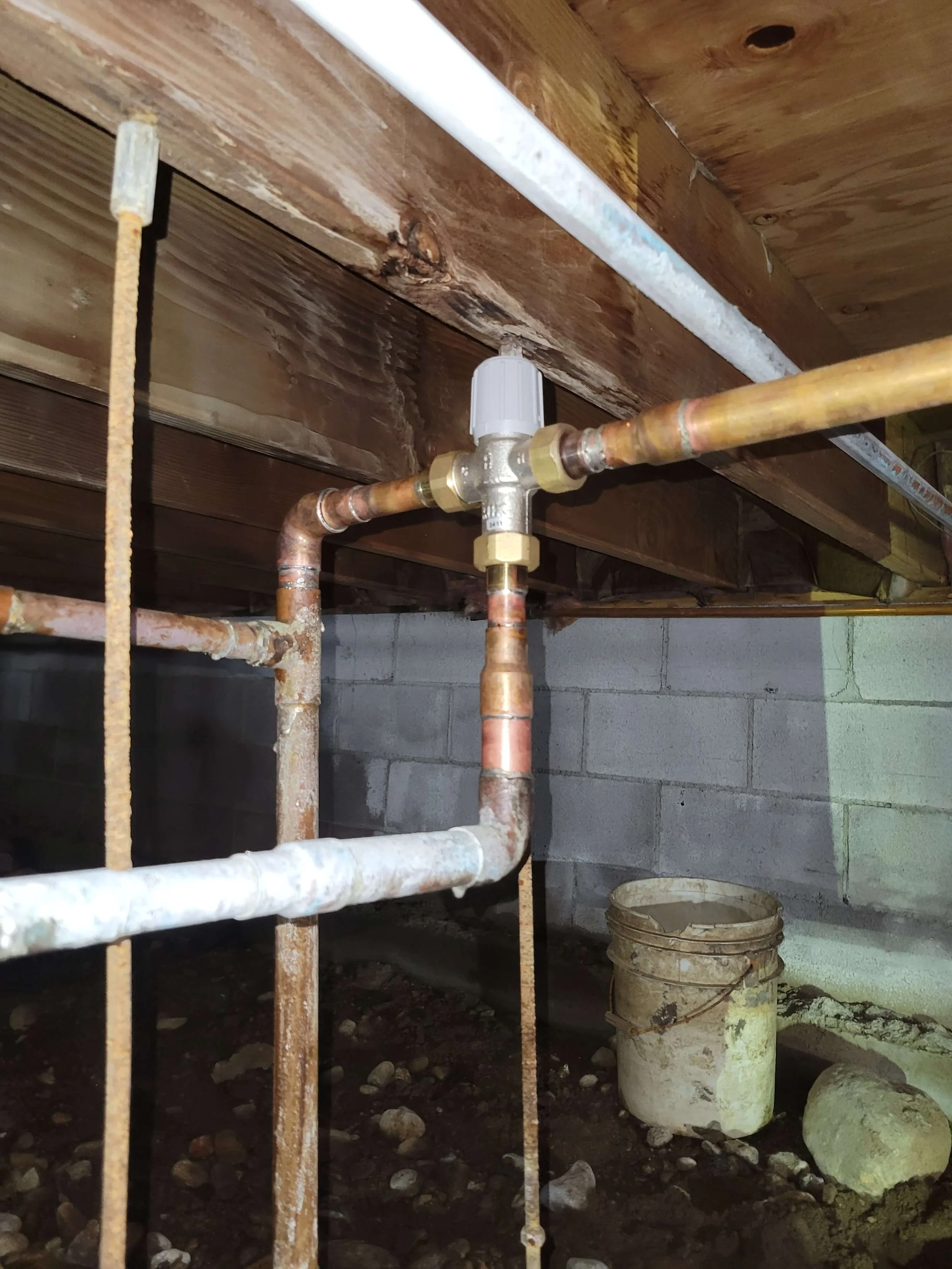 Crawl space showing plumbing system with pipes, fittings, and a pressure valve mounted under wooden beams, next to a concrete block wall and a dirty plastic bucket on the ground.