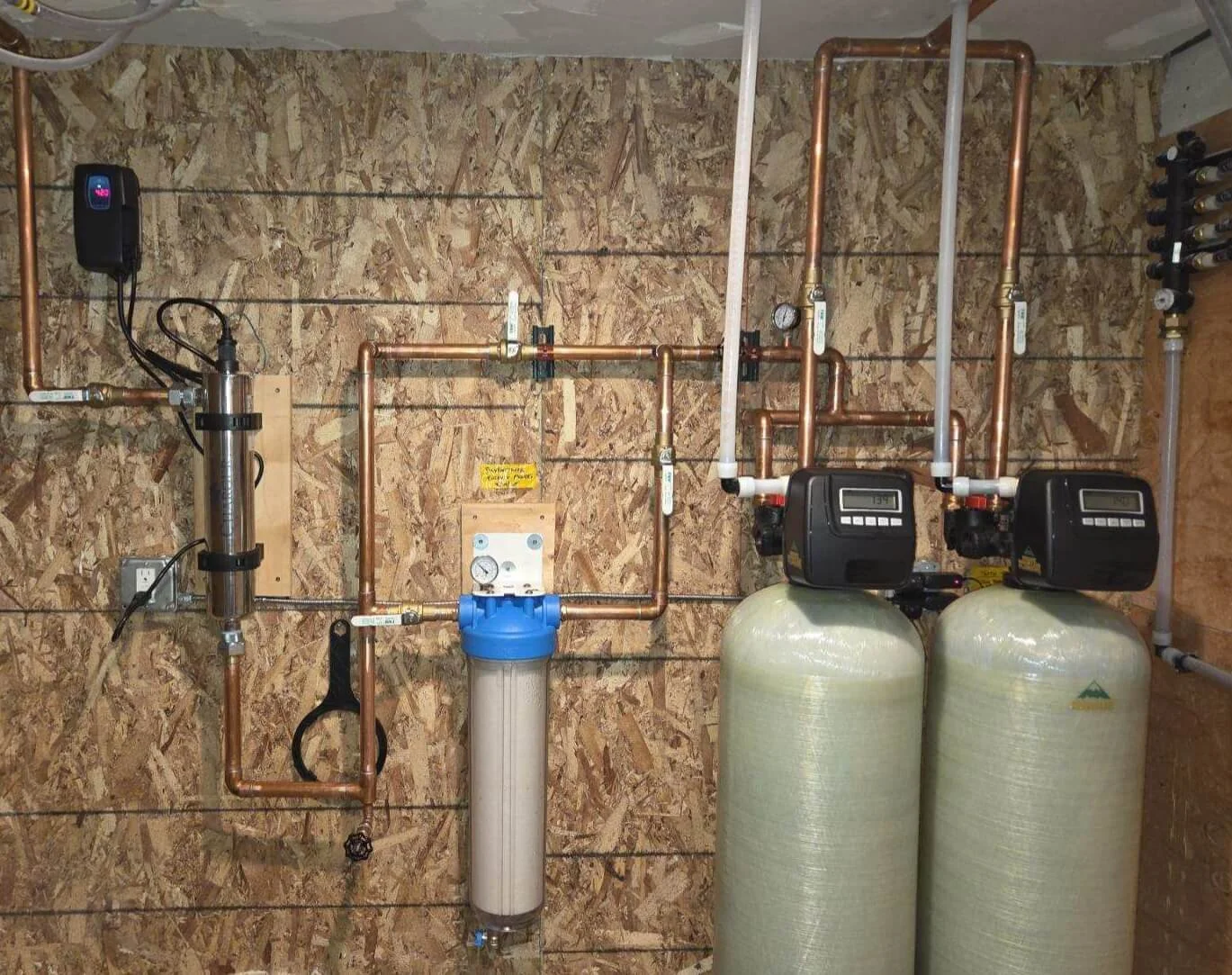 Plumbing system with copper pipes, water filters, and large water softener tanks against a wooden wall.