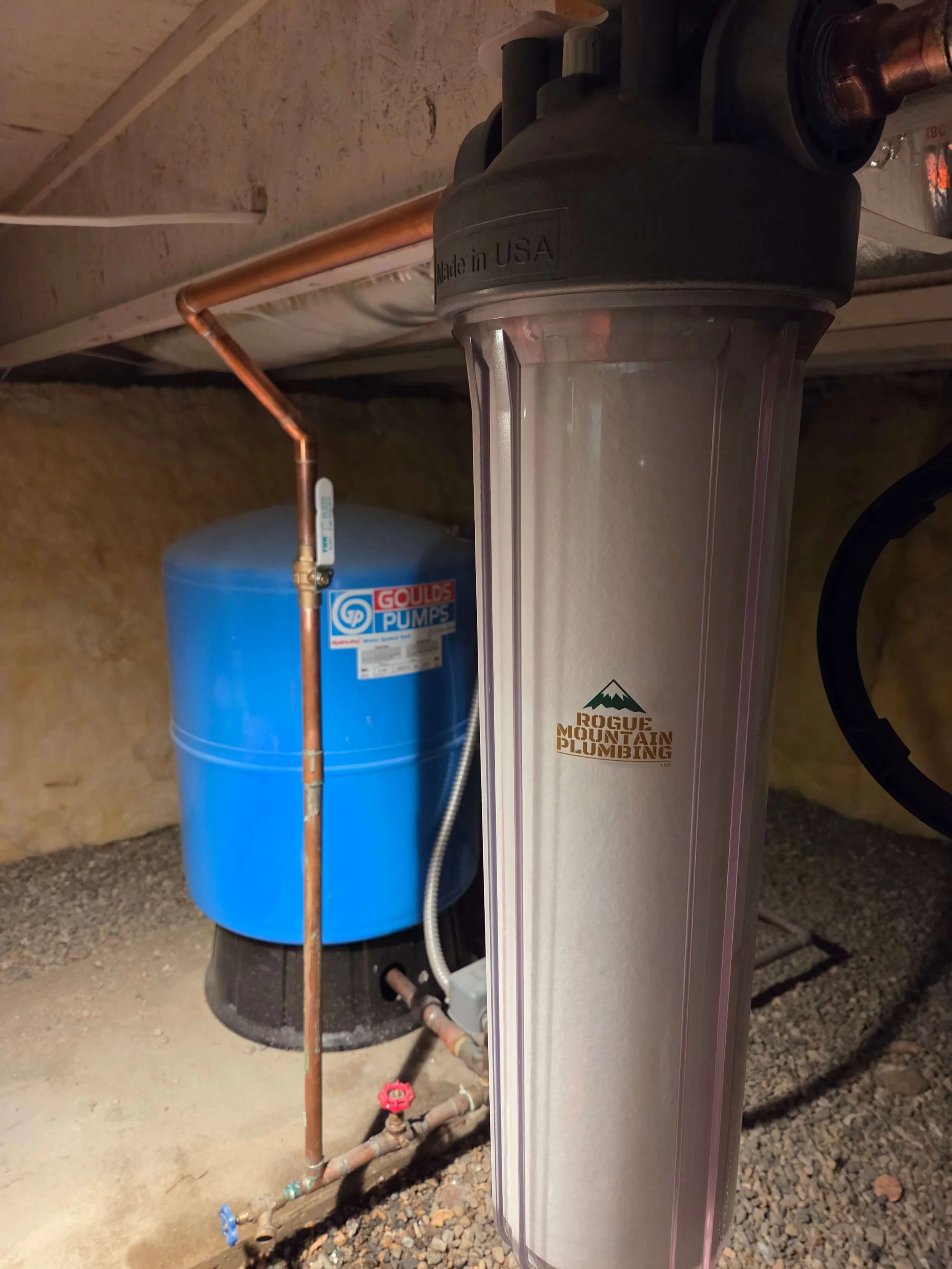 Water filtration system and pressure tank in a basement, with plumbing and gravel on the floor.