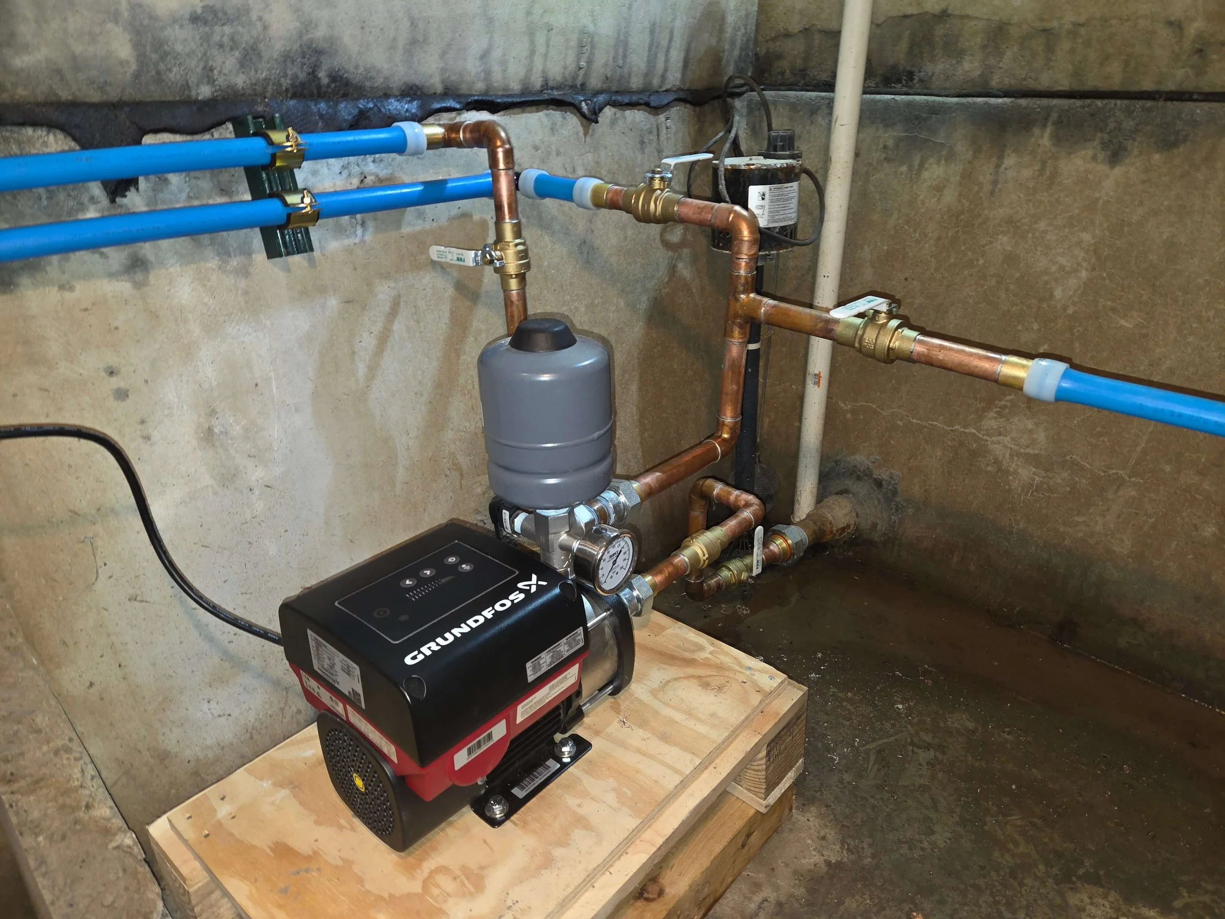 Pump and plumbing system with copper and blue pipes, a gray expansion tank, and a Grundfos pump unit installed on a wooden platform in a basement or utility room.