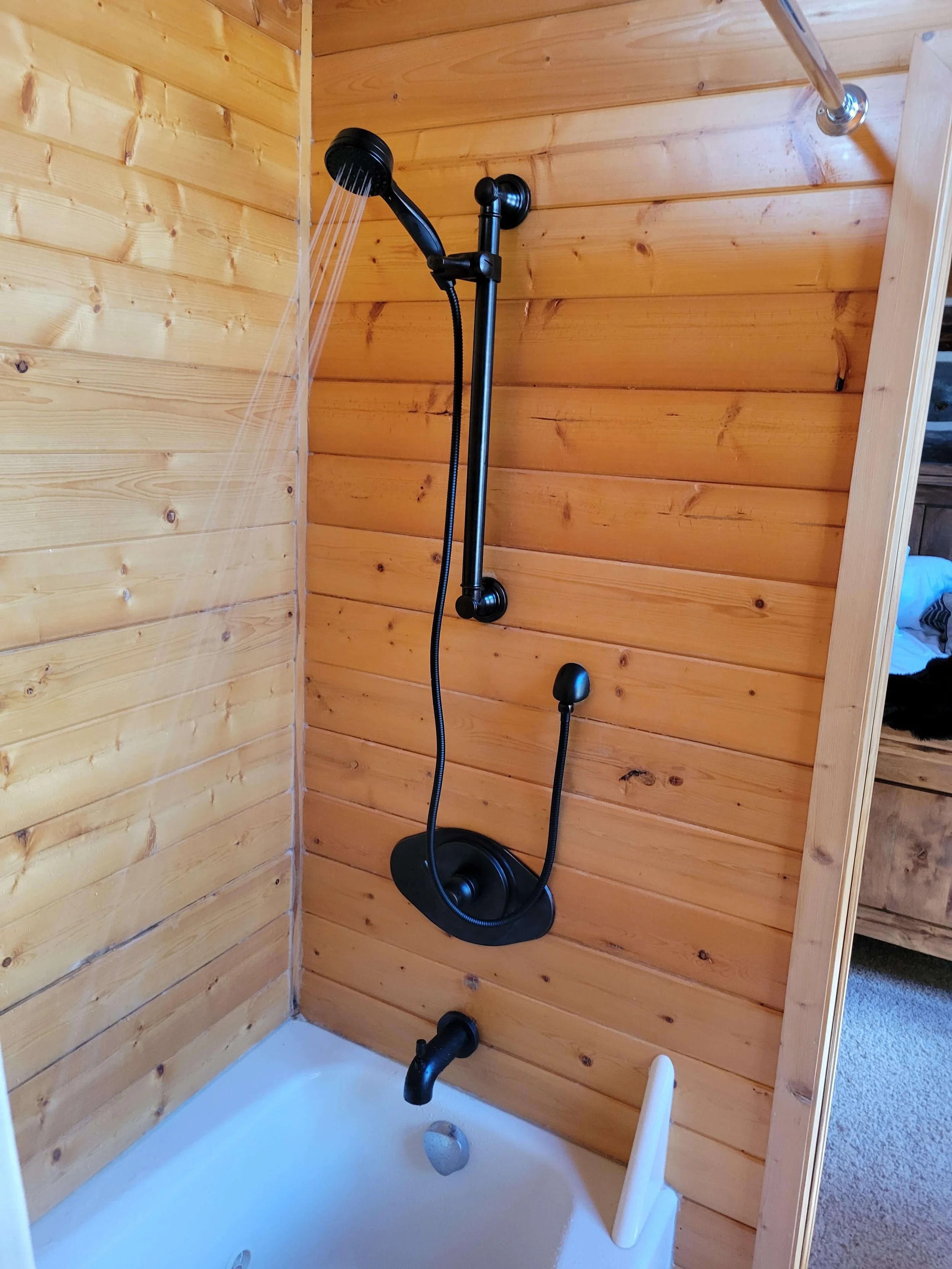 Wood-paneled bathroom with a showerhead and a small bathtub. The showerhead is attached to a wall-mounted bar, and water is flowing from it. The bathtub has a faucet and a drain. The walls have a natural wood finish.