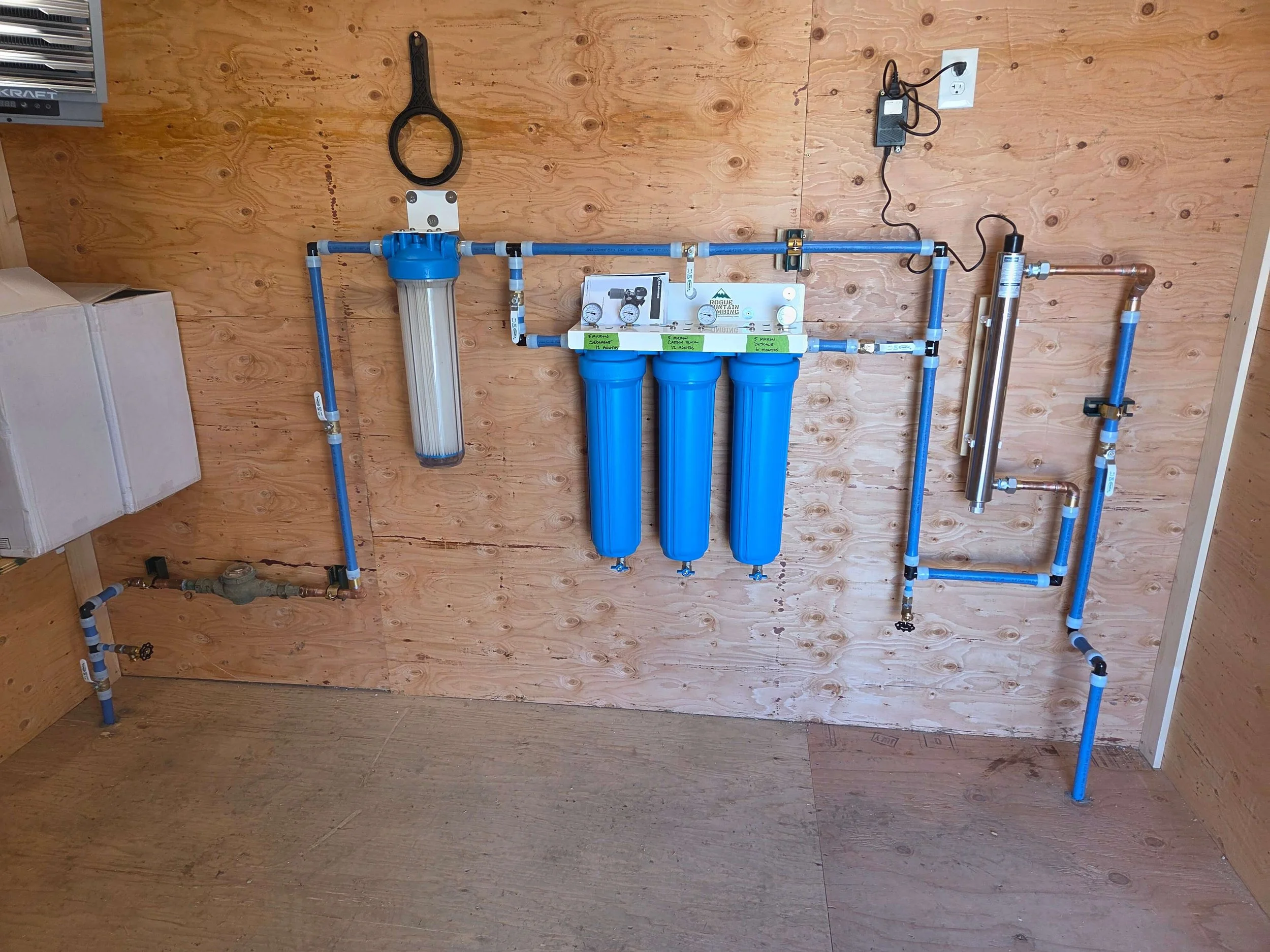 Water filtration system with three blue filter housings, connected blue piping, mounted on a wooden wall, with filtration equipment and electrical components.