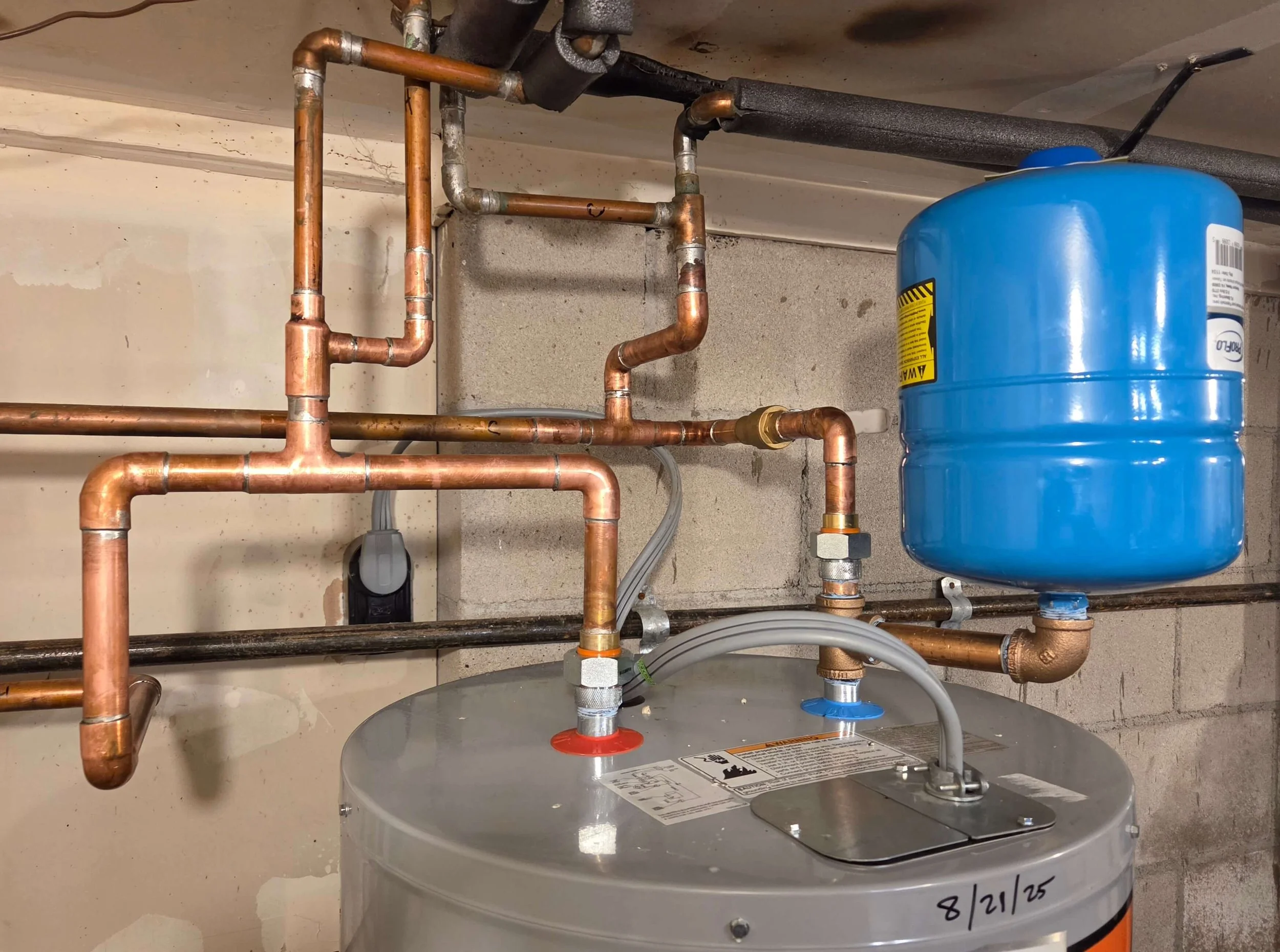 A water heater with copper pipes connected to it and a blue expansion tank mounted on the wall behind it.