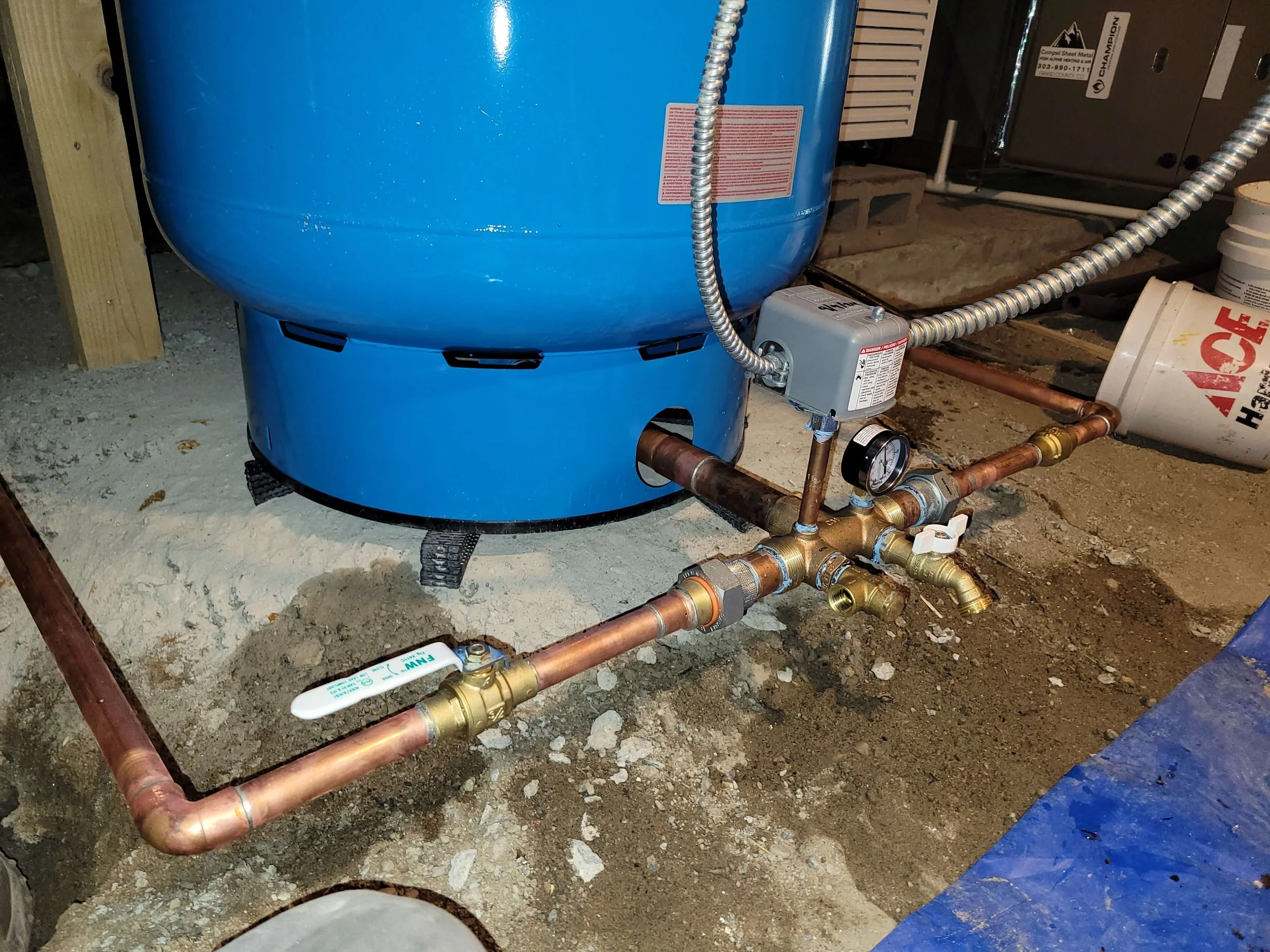 Blue water pressure tank connected to copper pipes and a pressure gauge, installed in a basement or utility area.