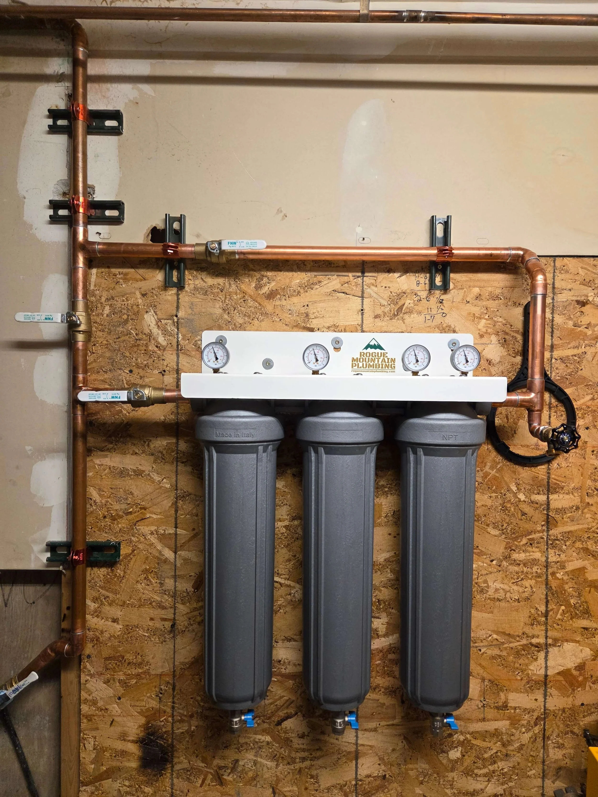Water filtration system with three large gray filter housings mounted on a wooden wall, connected by copper pipes with pressure gauges on top.