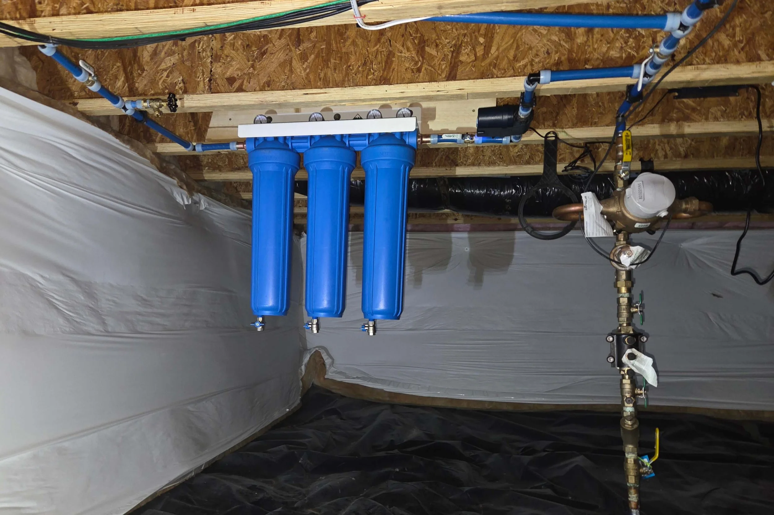 Plumbing system with three blue water filtration tanks, connected to pipes, valves, and a water meter on a wooden wall inside a building under construction.