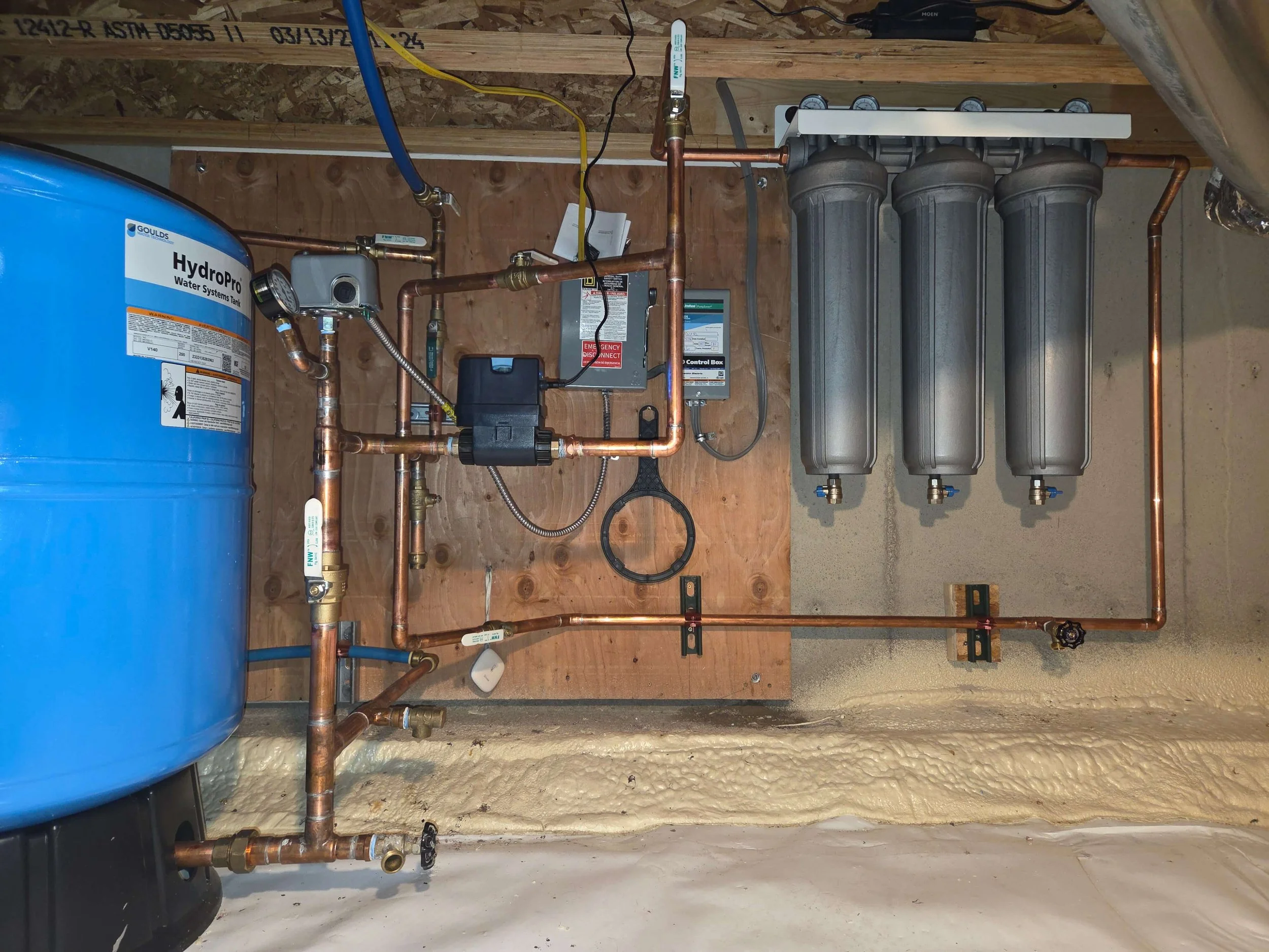 A water treatment system with a large blue pressure tank on the left, copper pipes, a control box, and three large metallic filters on the right, mounted on a wooden wall in a utility room.